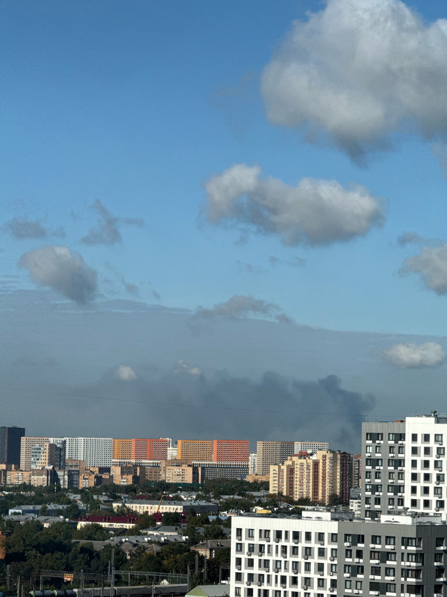 A high-angle, wide shot captures a residential area in Lyubertsy, Russia under a bright blue sky dotted with white clouds. In the foreground, modern, white and grey apartment buildings with many windows dominate the view. Behind them, a row of taller apartment buildings display a variety of colors, including orange, yellow, and white. Further in the background, a hazy plume of dark smoke rises into the sky, originating from an unseen source in the middle ground. The rest of the scene is filled with more apartment buildings and lush green trees. No people are visible in the frame.