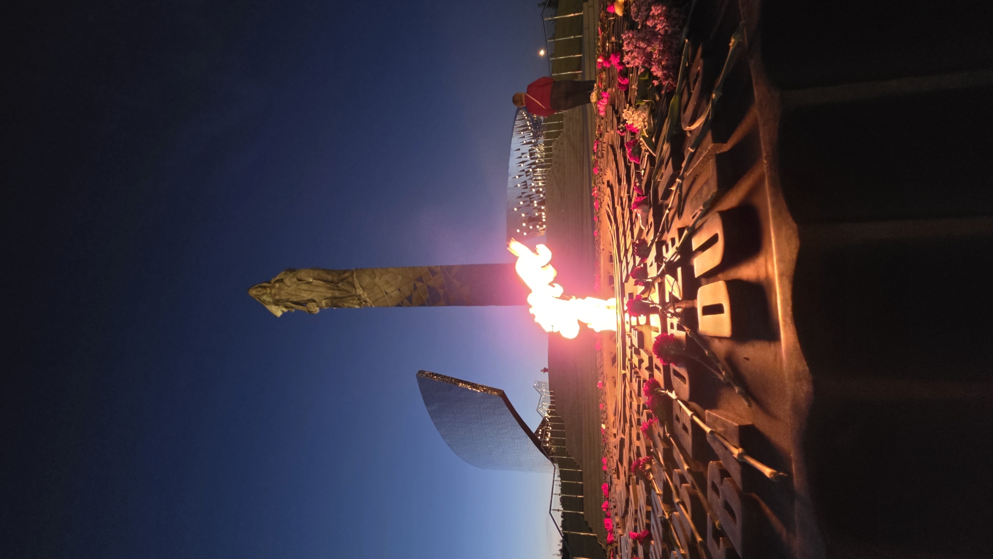 At dusk in an unidentified city, a memorial complex is visible. A vertical monument stands tall, topped with a sculpted figure. A flame burns brightly, appearing to emanate from a structure adorned with cut-out shapes and adorned with red flowers. A reflective, angled panel is positioned nearby. A person in a red shirt stands at the back of the area, possibly at the top of steps with railings. The sky transitions from dark to lighter blue.