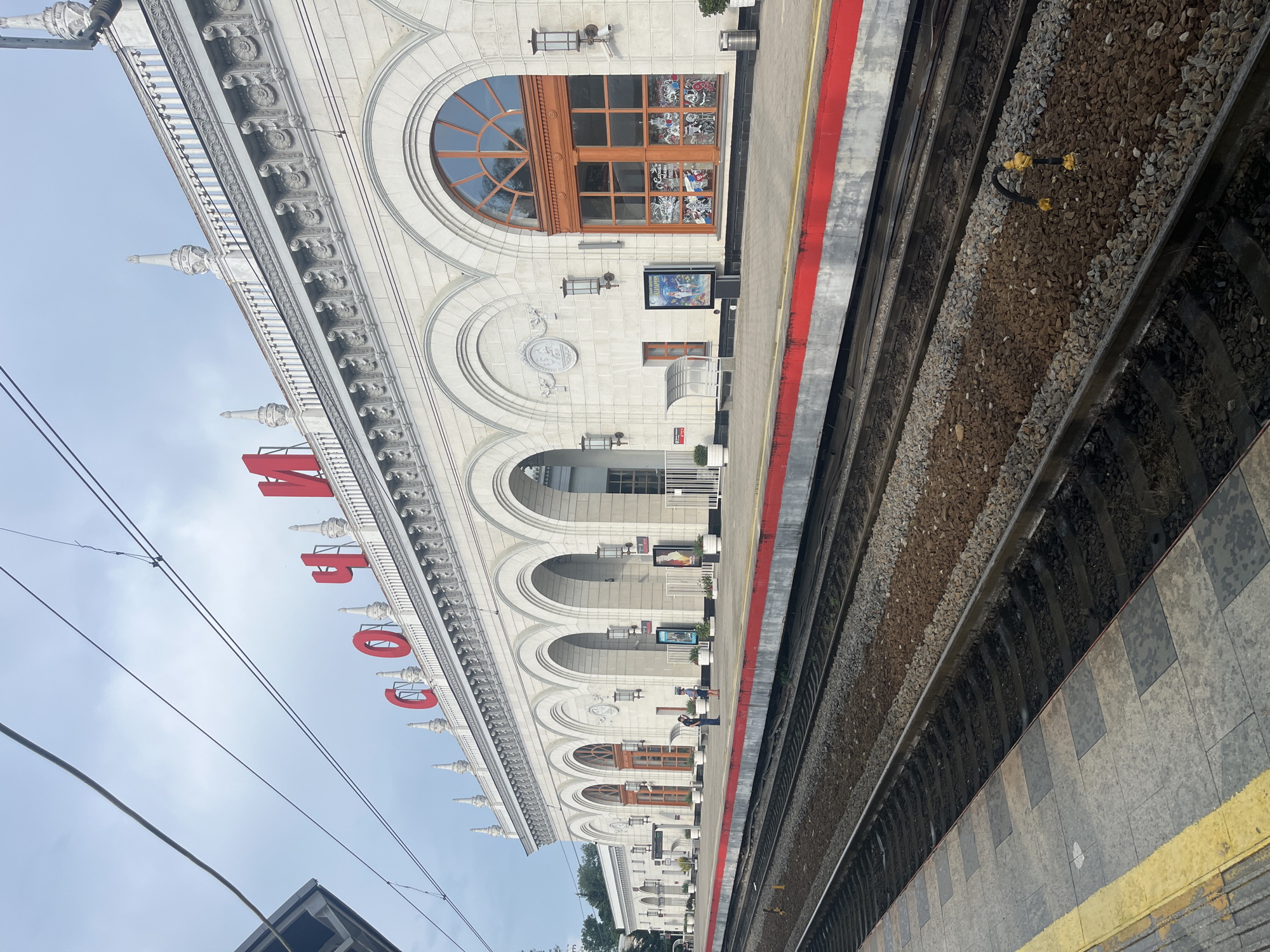 The image shows a view of the Sochi railway station in Sochi, Russia. The station building is grand, with a white facade adorned with arches and ornate detailing. Red lettering spelling "СОЧИ" (Sochi) is prominently displayed on the roofline. The platform in the foreground has a red stripe along its edge. Several sets of train tracks are visible, with gravel ballast between them. Two people are standing on the platform, appearing to be waiting. Overhead, electric train wires are strung across the sky, and the sky is overcast. Posters are displayed on the building facade, and potted plants add greenery to the platform area.