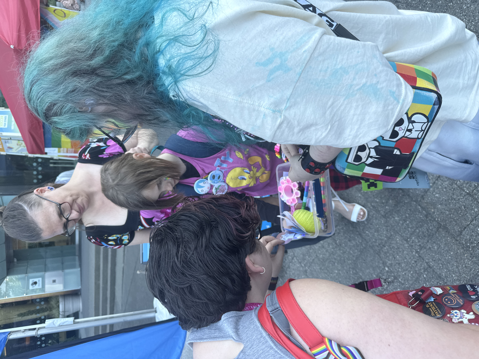 Four people are gathered around a tray of craft supplies. One person, on the left, with short dark hair and a grey tank top with red straps, is leaning down and looking at the tray. Another person, in the center, with brown hair and wearing a pink t-shirt adorned with Tweety Bird and various pins, is also looking at the tray. To the right, a person with long, wavy teal hair and glasses is wearing a cream-colored t-shirt and a multi-colored bag decorated with cartoon figures. In the background, another person wearing glasses and a black and pink patterned shirt is observing the group. The scene appears to be outdoors, possibly at a community event or festival, with a blue tent on the left and a red awning with rainbow detailing visible in the upper right corner. The ground is paved. The overall impression is one of engagement and shared activity.