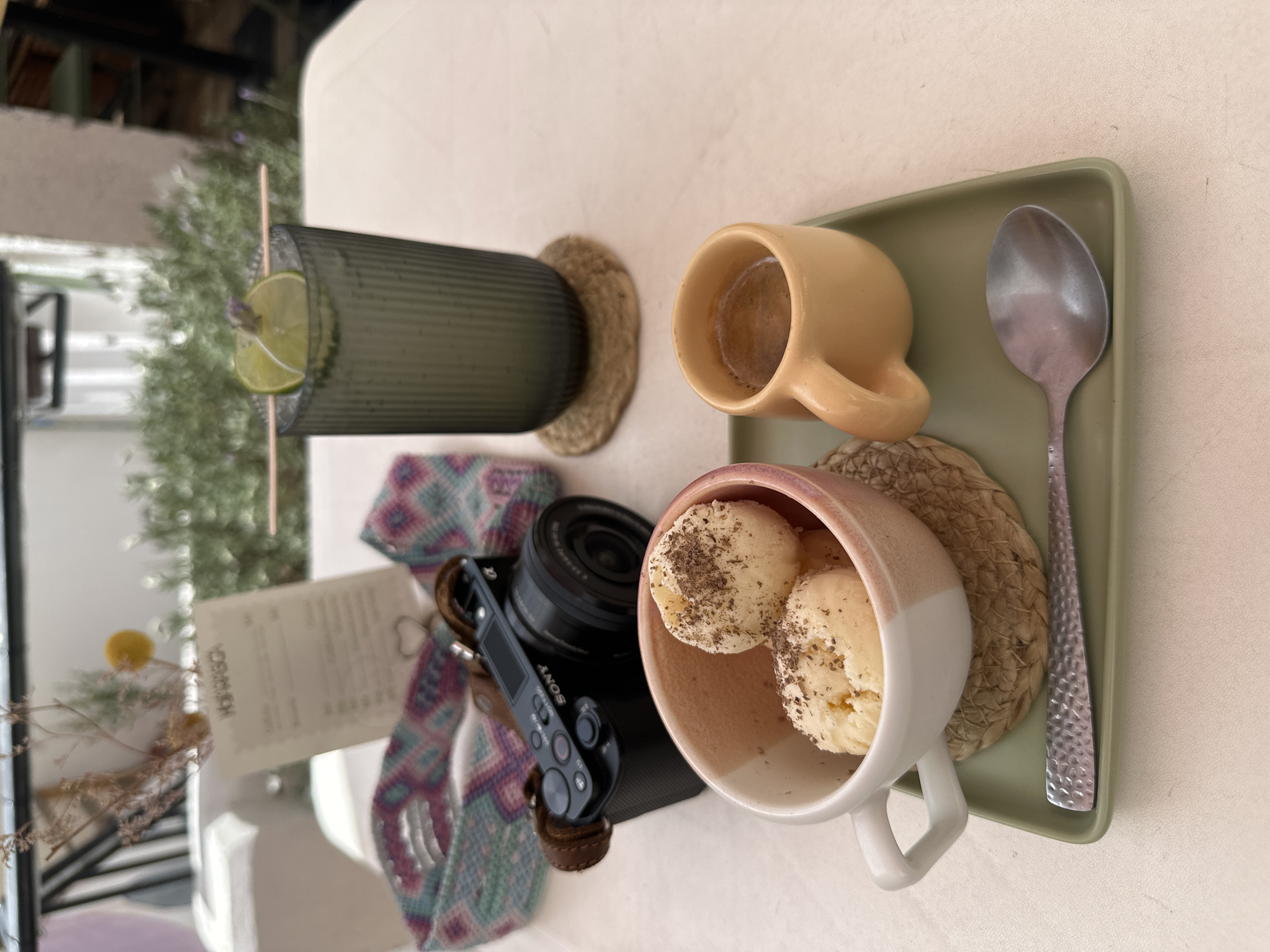 A table in an outdoor cafe in Zacatecas City, Mexico, displays a spread of food and drink. A Sony camera with a leather strap rests on the table near a decorative woven band. In the foreground, two scoops of ice cream are served in a pink-rimmed white cup atop a woven coaster. Beside the ice cream is a small yellow mug containing a dark liquid, likely coffee. A silver spoon lies on a green tray in front of the cups. In the background, a tall glass with a lime and lavender garnish contains a beverage, and a menu is propped up behind the camera. The setting suggests a relaxed outdoor dining experience.