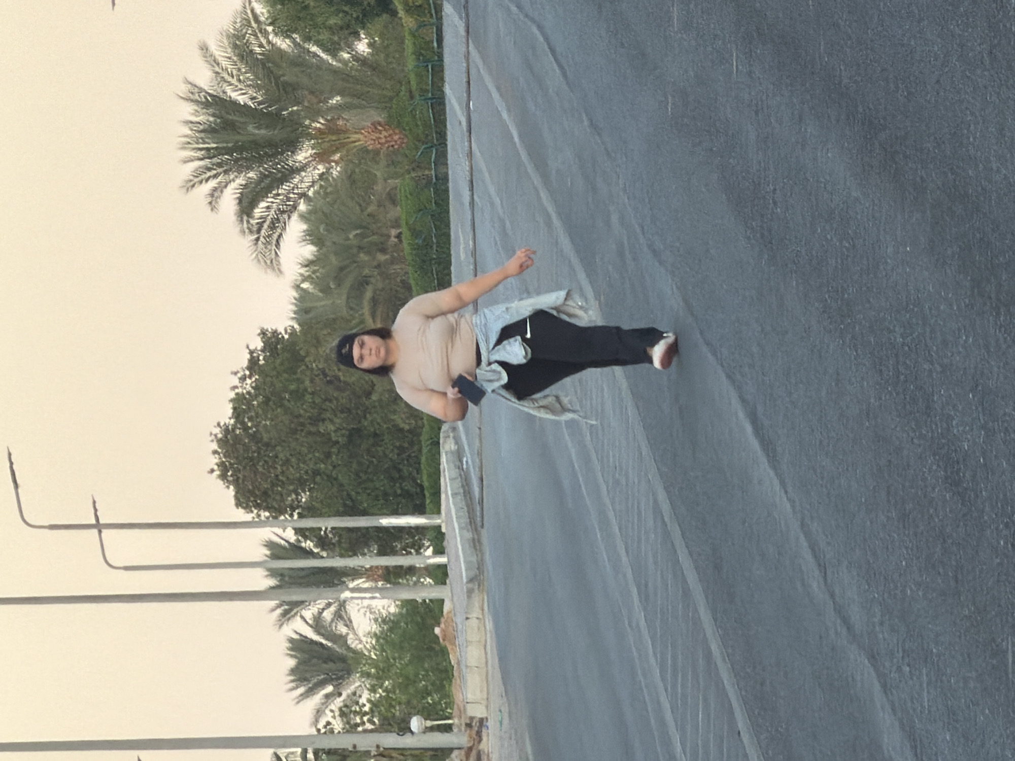A full-shot, eye-level, outdoor, slightly blurry, medium-distance photograph captures a woman walking towards the camera on a paved road with multiple lanes. She is wearing a light brown t-shirt, black pants with a white stripe, and a light denim jacket tied around her waist. She also wears a black baseball cap and white sneakers with red accents. She is holding a black smartphone in her left hand.
The background reveals a street with palm trees and other vegetation, characteristic of a tropical or subtropical environment. Several tall streetlights are visible in the background, along with a light-colored sky. The road appears to be a public thoroughfare in Sharm El Sheikh, Egypt, possibly a promenade or main road leading into the city, given the presence of streetlights and the style of vegetation. The lighting suggests it is either early morning or late afternoon.