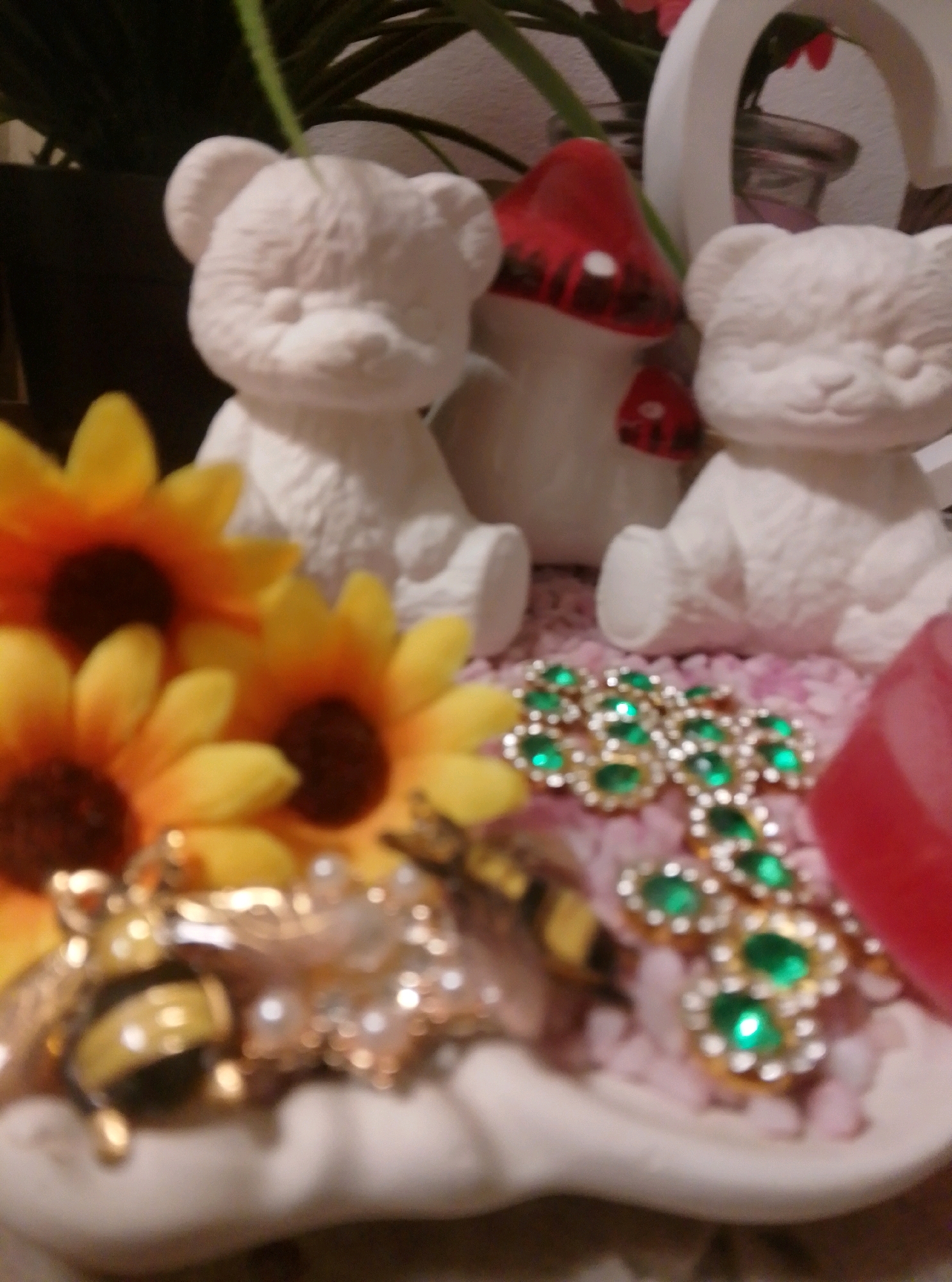 This is a close-up, slightly out-of-focus, indoor photograph of decorative items. In the foreground, there are several artificial sunflowers with yellow petals and dark centers. Partially visible behind the sunflowers are two white, unpainted ceramic teddy bear figurines and a red and white mushroom figurine. A cluster of gold-toned brooches with green gemstones and small white pearls is arranged on the surface. A decorative bumblebee charm is also visible amongst the brooches. The items are resting on a white, shell-shaped dish filled with pink decorative filler. A portion of a red object, possibly another decorative item, is visible on the right side of the image. The background is blurred, showing dark green foliage and part of a white arch-shaped object. The image does not contain any discernible location information for the city of Wedel, Germany.