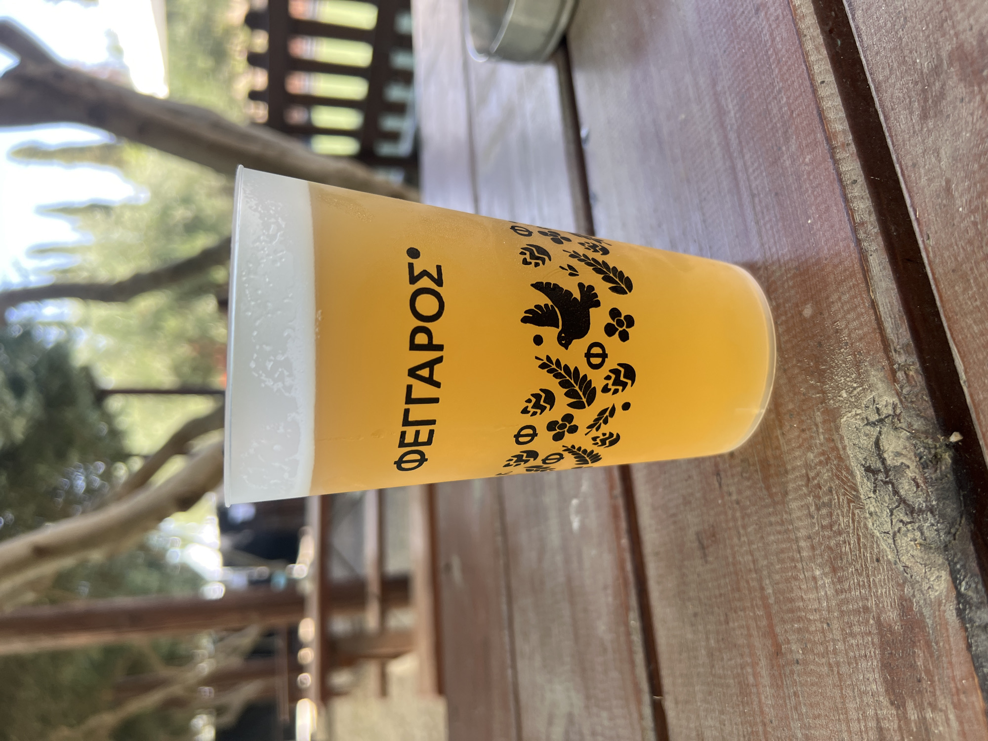 A close-up shot shows a pint glass filled with a golden-hued beverage, topped with a frothy white head, resting on a weathered wooden table. The glass is adorned with black graphics, featuring the word "ΦΕΓΓΑΡΟΣ" in Greek lettering at the top, followed by a decorative pattern of leaves, flowers, and a bird. The background, out of focus, reveals an outdoor setting with trees, foliage, and what appears to be a wooden fence or railing. The overall impression is that of a relaxed, possibly outdoor dining or drinking establishment, likely in a rural or semi-rural location. The specific location in Analiontas, Cyprus is inferred from the context provided, suggesting a natural or agricultural environment.