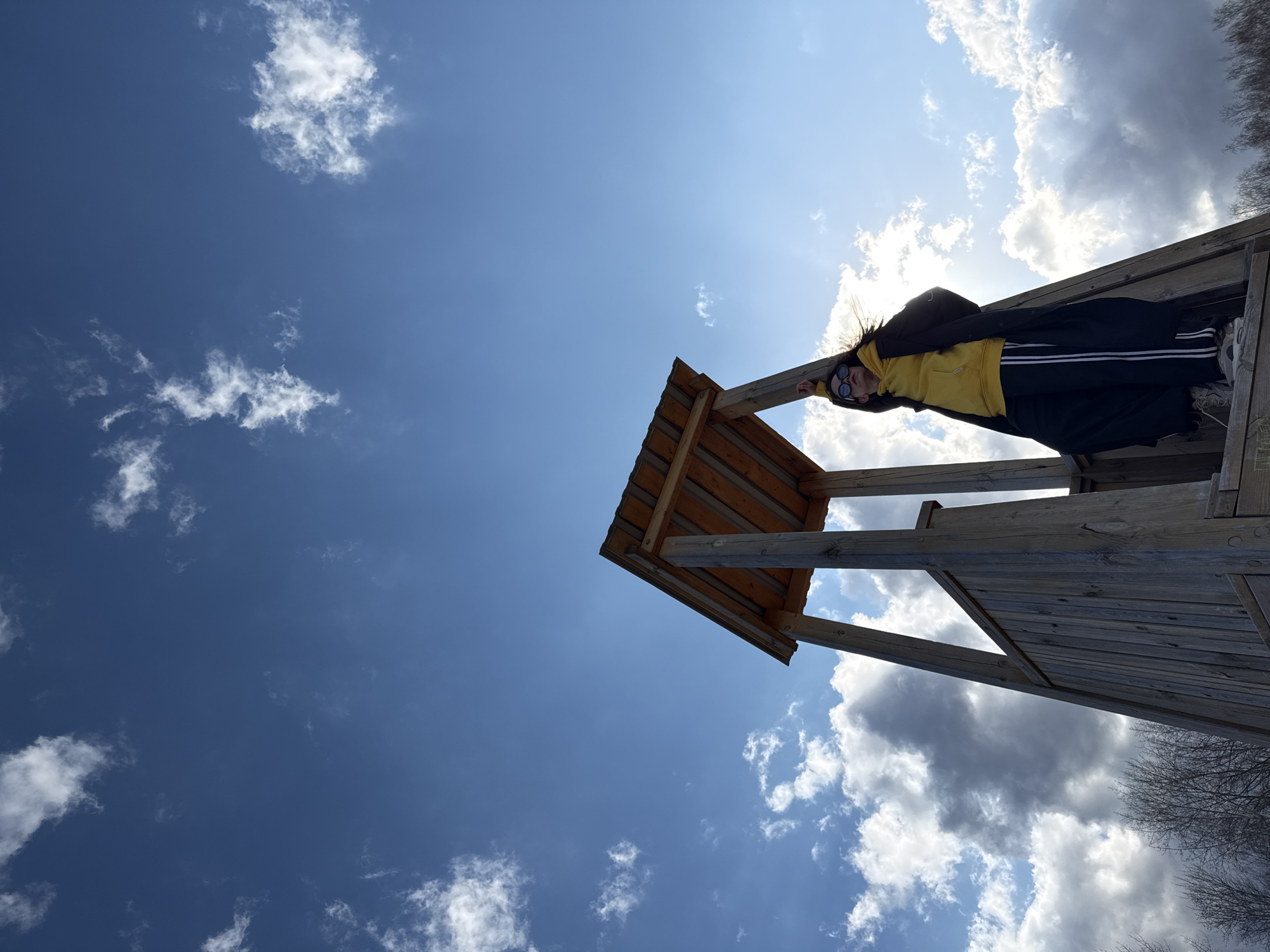 A low-angle, wide shot captures a person on a wooden structure against a bright blue sky with scattered white clouds. The image is rotated, giving the impression the person is lying horizontally or at an extreme angle, looking down towards the viewer. The sunlight is directly behind the person, creating a strong backlight and some lens flare, highlighting the edges of their hair.
The main subject is a person wearing a yellow hoodie, a dark jacket, dark pants with white stripes down the side, and sneakers. They are also wearing sunglasses and have long, dark hair which appears windblown around their head. The person is positioned on a rustic wooden structure, possibly an observation deck, a tower, or a raised platform, with visible wooden beams and planking.
The scene is outdoors during bright daylight. The weather is sunny with a mix of clear sky and fluffy white clouds. In the far right background, the silhouettes of bare tree branches are visible, suggesting a season like late winter or early spring in a location such as Chaikovsky, Russia. There is no visible text in the image. The pose appears casual or playful, possibly taken from an unusual perspective to create a dynamic effect.