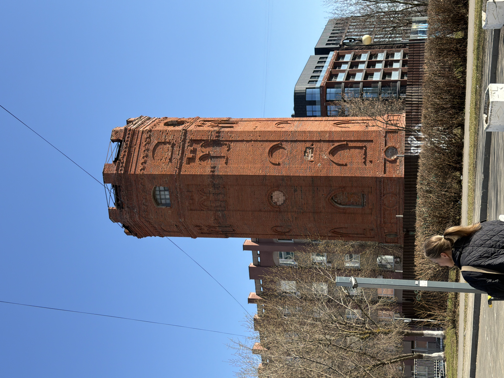 The image shows the brick water tower on Karl Marx Street in Izhevsk, Russia. The tower is a prominent structure, standing tall against a clear blue sky. Details of the surrounding urban environment are visible, including nearby buildings, trees, and a sidewalk. A woman is standing near a street lamp, she is dressed in a dark quilted jacket with blonde hair pulled back in a ponytail.