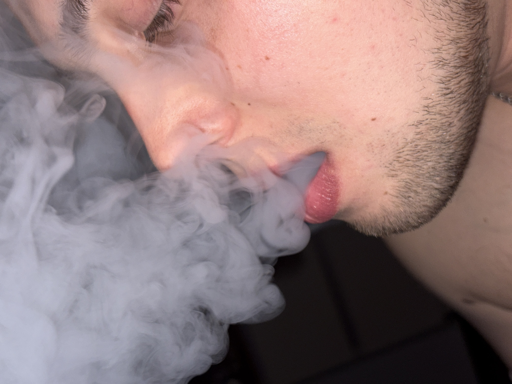 The image shows a close-up of a person exhaling a large plume of white vapor. The person has light skin, dark hair, and a trimmed beard. The vapor is concentrated around the mouth and dissipates outwards. The background is dark and out of focus. The city and exact location are unknown.