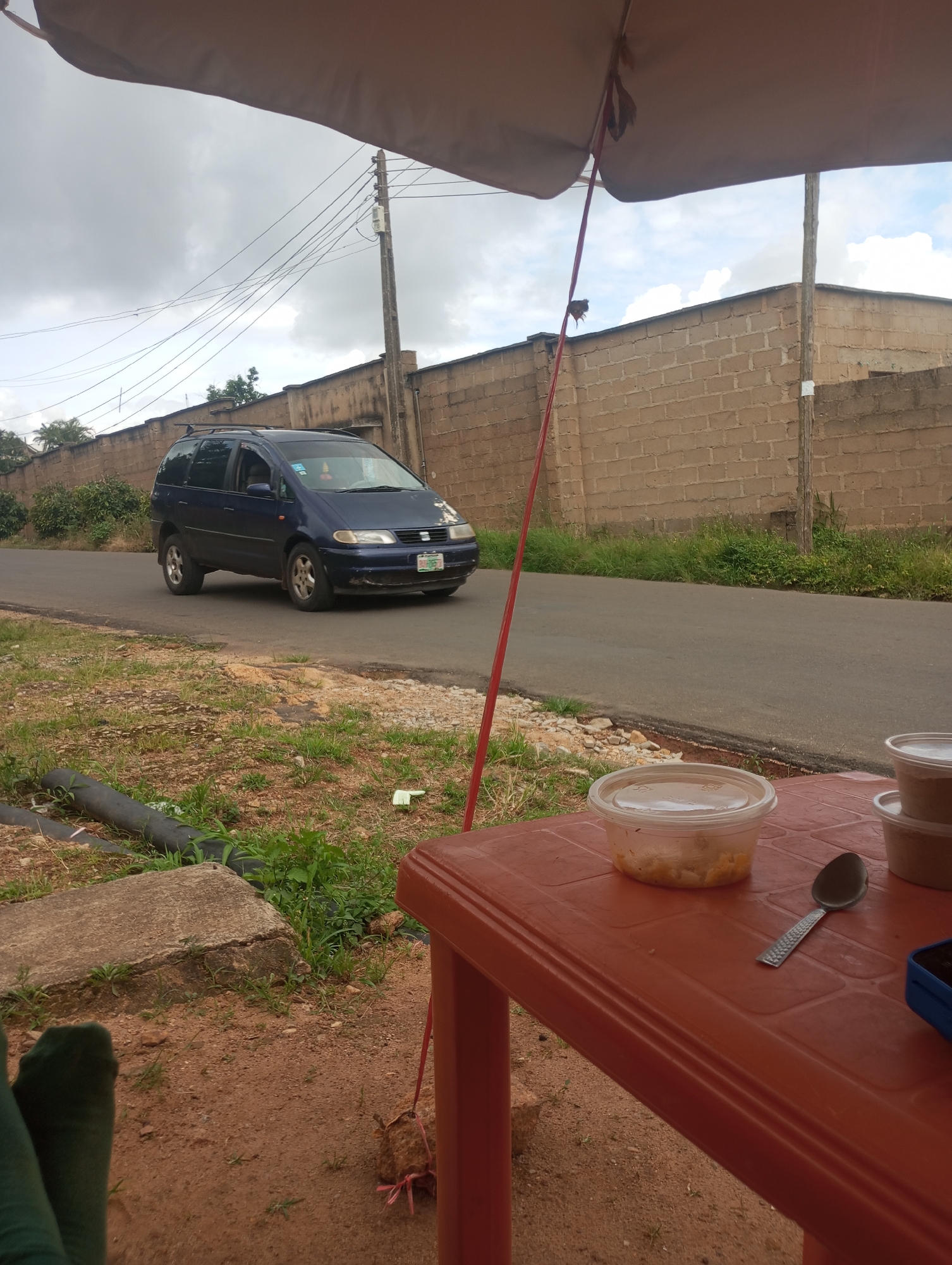 In Jos, Nigeria, a blue minivan drives down a paved road bordered by a brick wall and overgrown grass. In the foreground, a reddish-brown plastic table holds two takeout food containers and a spoon. The table is situated on a patch of sandy ground with some scattered debris, including two pieces of black plastic pipe. The scene is covered by a large tan awning, casting a shadow over the table and food. The sky is overcast with a few white clouds. The overall environment appears to be a roadside setting, possibly near a residential or commercial area.