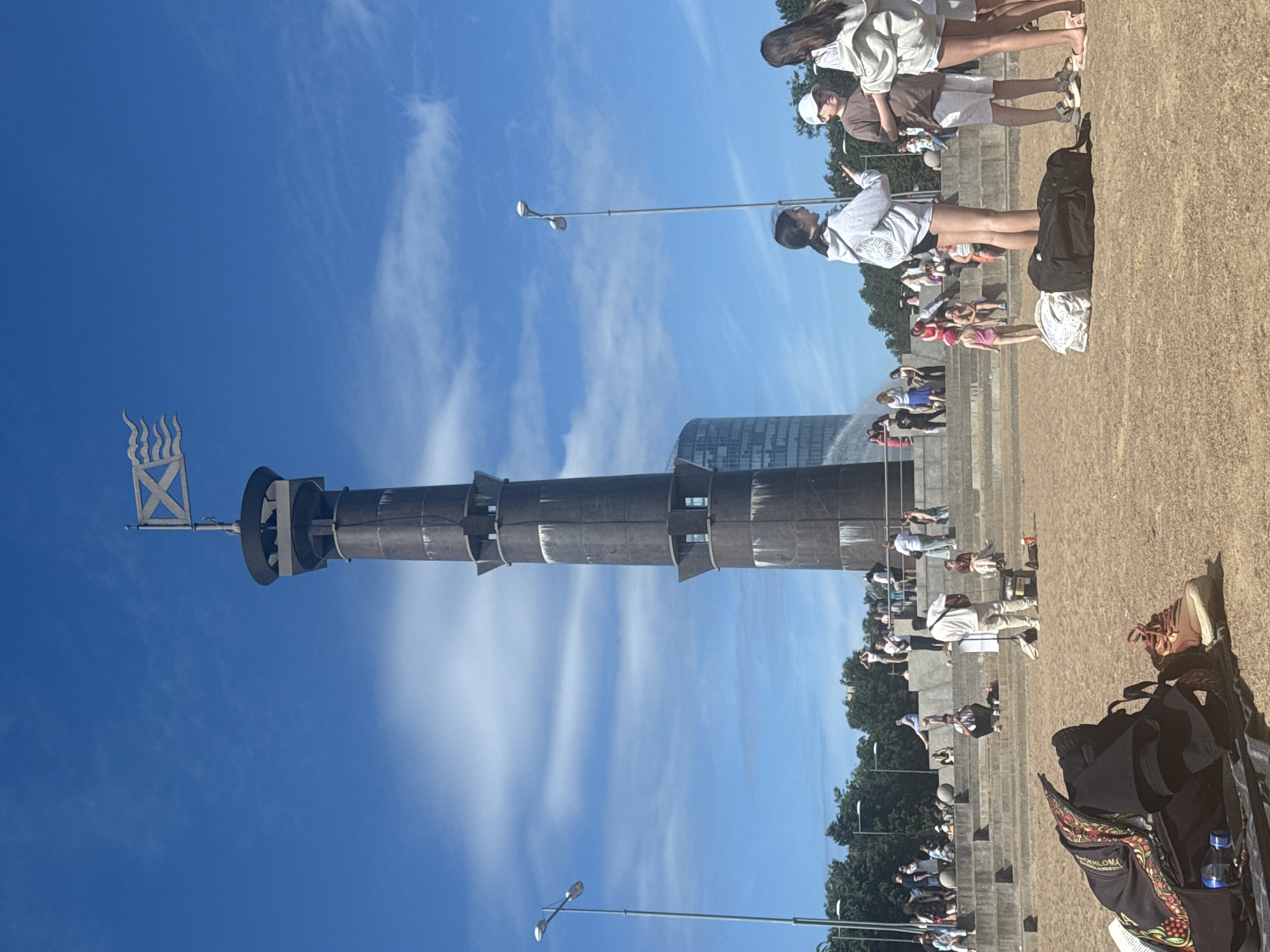 The image depicts a sunny day in Saint Petersburg, Russia, near the Monument to the Founders of St. Petersburg on the Strelka of Vasilyevsky Island. In the foreground, several people are seen gathered on a sandy area, with backpacks and bags placed on the ground. One backpack in the lower left corner displays a colorful, intricate pattern.
In the mid-ground, a tiered concrete structure serves as a viewing platform where a crowd of people are seated and standing. Some individuals are engaged in conversation, while others appear to be observing the surroundings. A man in a white outfit is playing a guitar, drawing the attention of some onlookers.
Dominating the background is the Monument to the Founders of St. Petersburg, a tall, dark, cylindrical structure topped with a flag featuring a saltire and wavy lines. To the left of the monument, a modern glass building is visible. The sky is a clear blue with scattered white clouds. The overall scene suggests a relaxed public gathering in an urban outdoor space.