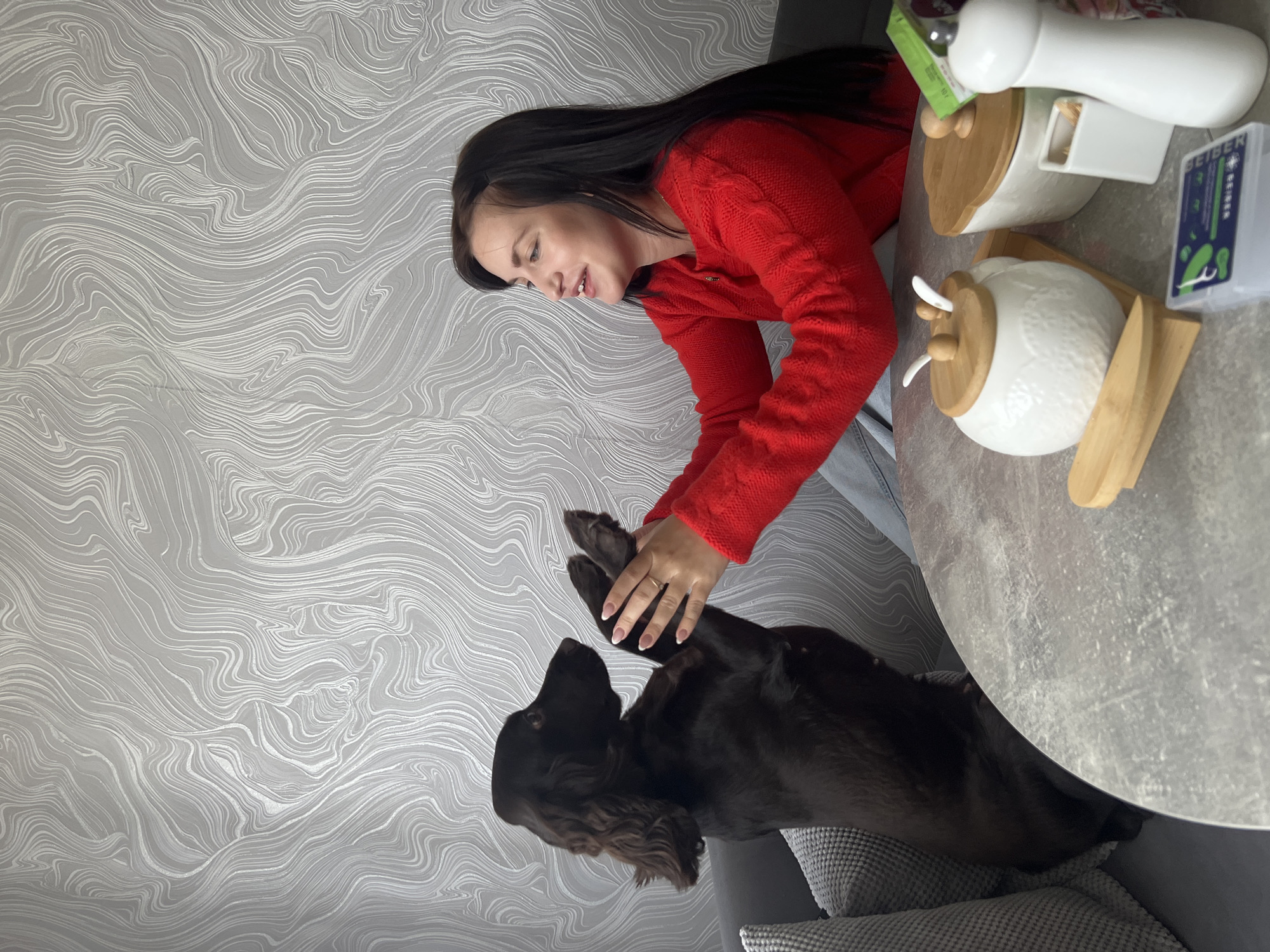 The image shows a woman with long dark hair wearing a red sweater. She is interacting with a brown spaniel-type dog by holding its paws. The dog is standing on a gray upholstered sofa. A round gray table is partially visible on the right. On the table are two white ceramic containers with wooden lids and a white utensil holder with a white utensil inside. The wallpaper is gray with a white swirling pattern. The photo was taken indoors in Марковский, Russia.
