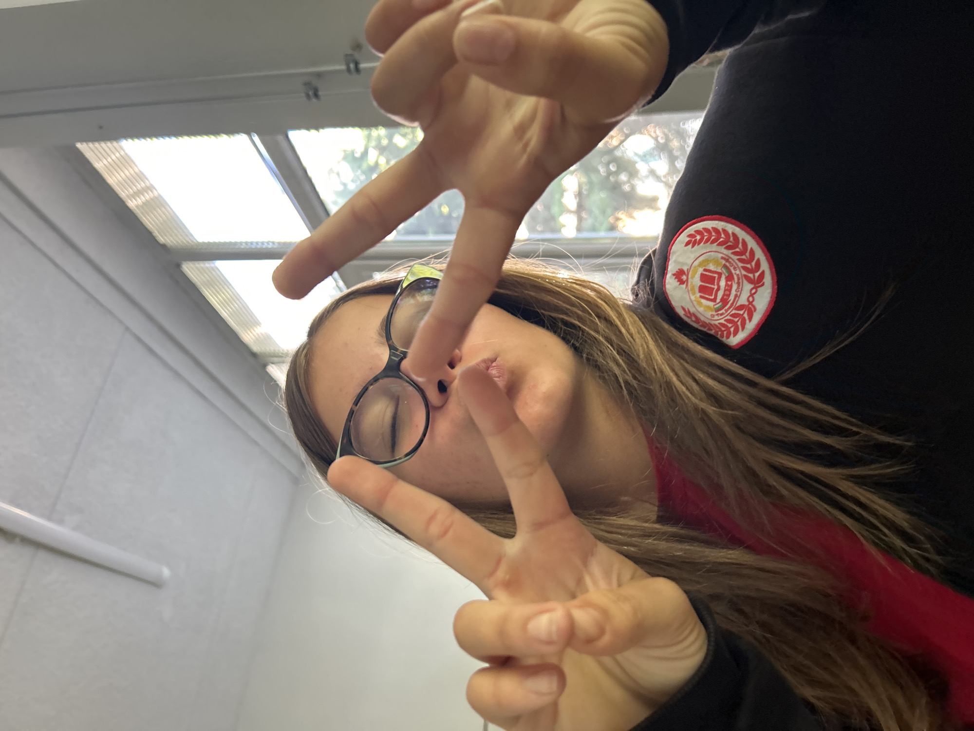 A close-up, angled shot captures a person, likely a young female, posing indoors. She has long, light brown hair and wears clear-rimmed glasses, which are slightly askew on her nose. Her face is largely obscured by her hands, both of which are held up in a "peace sign" or V-gesture, creating a playful or casual mood. She is wearing a black long-sleeved top, with a red garment visible underneath, near her shoulder and neck.
On the upper arm of her black top, a prominent red and white circular patch is visible. The patch features a laurel wreath design surrounding a central emblem that appears to be a stylized fort or building. Text within the patch, written in Cyrillic, clearly reads "ОБЩИНА ЛОВЕЧ" (Municipality Lovech), identifying the patch as belonging to the Bulgarian city of Lovech, which aligns with the given location context of Sofia, Bulgaria.
The background suggests an indoor setting, possibly a classroom or office, with bright fluorescent lighting panels visible on the ceiling. A window offers a view of trees and foliage outside, indicating it is daytime. The walls are a plain light color, likely white or off-white, and a white rail or pipe is visible on the lower left.