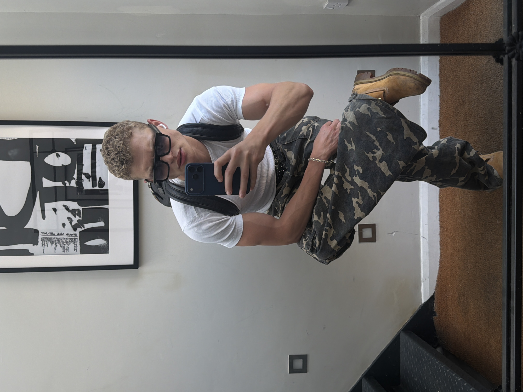 A young adult male poses for a mirror selfie indoors. He wears a white t-shirt, camouflage pants, tan work boots, and black sunglasses. He holds a smartphone in his right hand, capturing the reflection. A black messenger bag strap is visible across his chest. A black-framed abstract art print hangs on the wall to his left. A brown doormat and a metal railing appear in the background, suggesting an entryway or hallway. The location is inferred to be London, United Kingdom, based on the style of architecture and interior design elements. The man stands near a staircase, indicated by the glimpse of steps in the lower right corner.