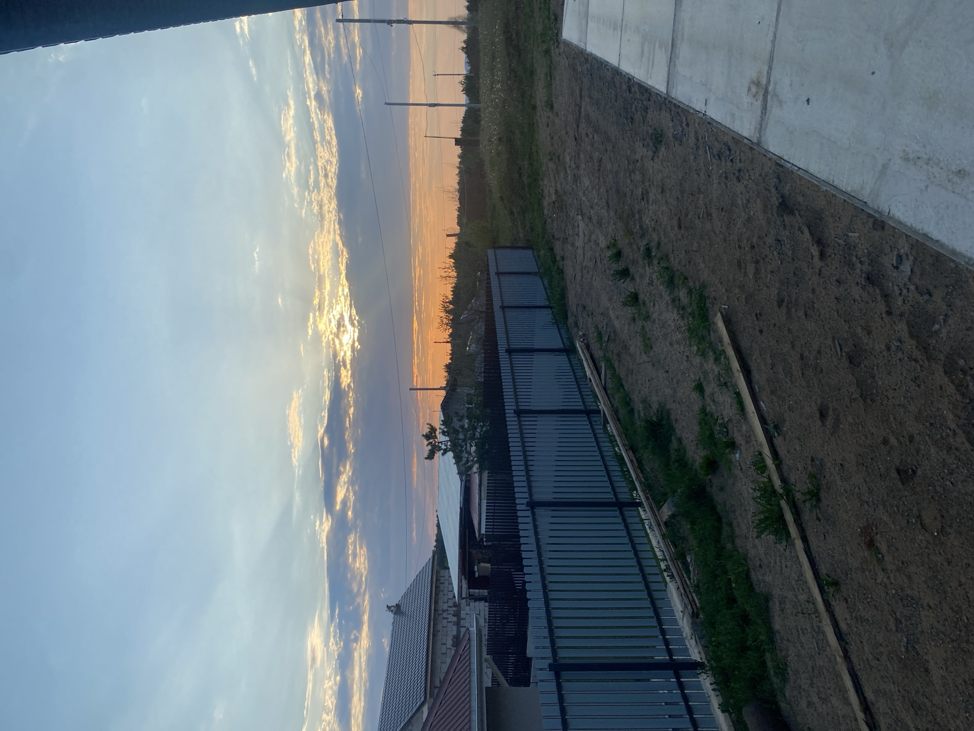 The image depicts an outdoor scene in what appears to be a residential area. The sky is visible, exhibiting a sunset with a gradient of blue merging into orange hues. Clouds are scattered across the sky, reflecting the light. Below the sky, a row of trees and power lines stretches into the distance.
In the foreground, a house with a pitched roof is visible, along with a dark metal fence. Next to the fence is an area of dirt and sparse vegetation. Adjacent to this, a concrete structure is present.