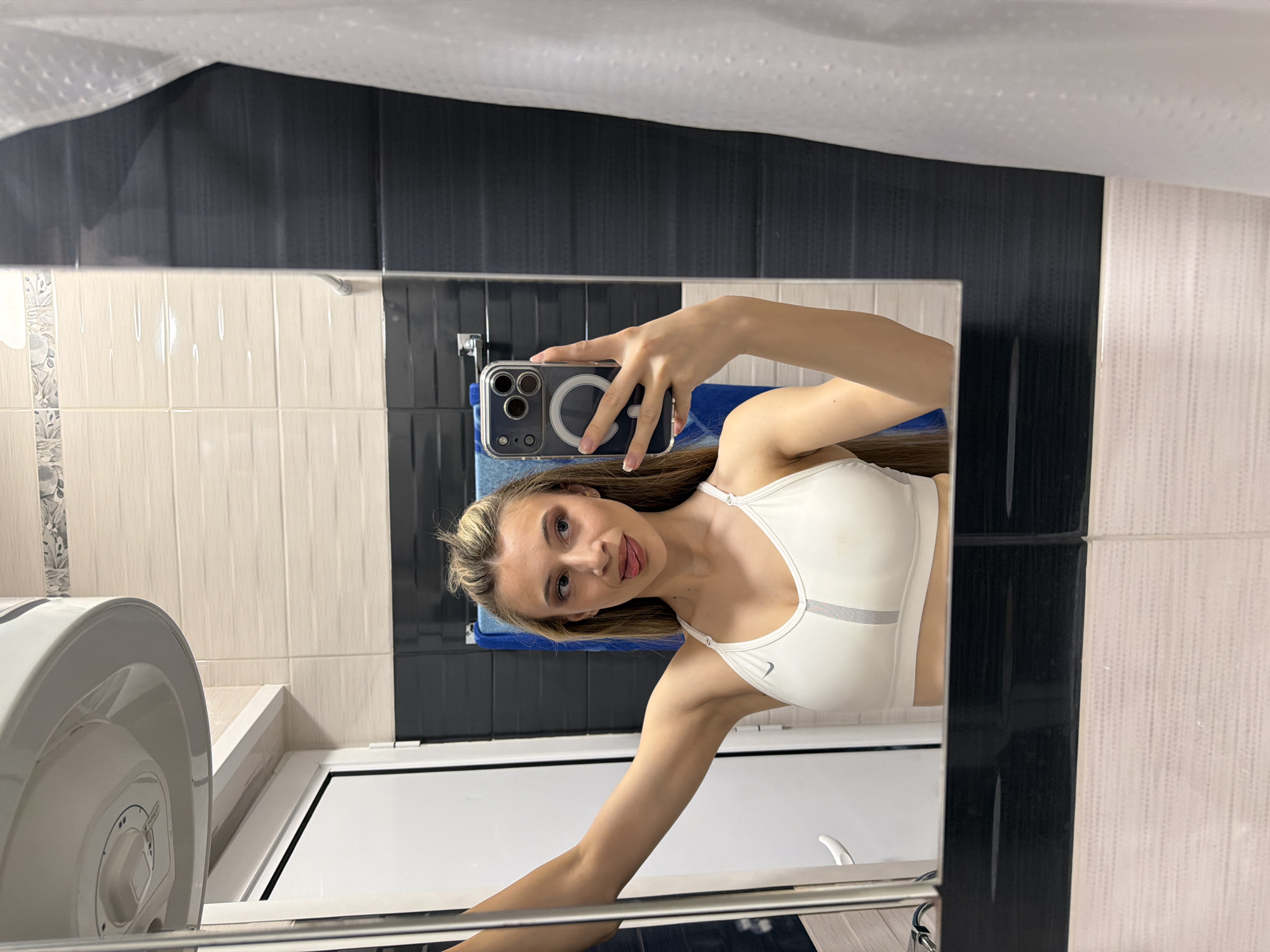A young woman is taking a selfie in a bathroom. She is wearing a white sports bra. She is holding a cell phone with a clear case. The bathroom has light beige and black tiled walls. There is a water heater on the left. A blue towel is visible on a rack. The location is Plovdiv, Bulgaria.