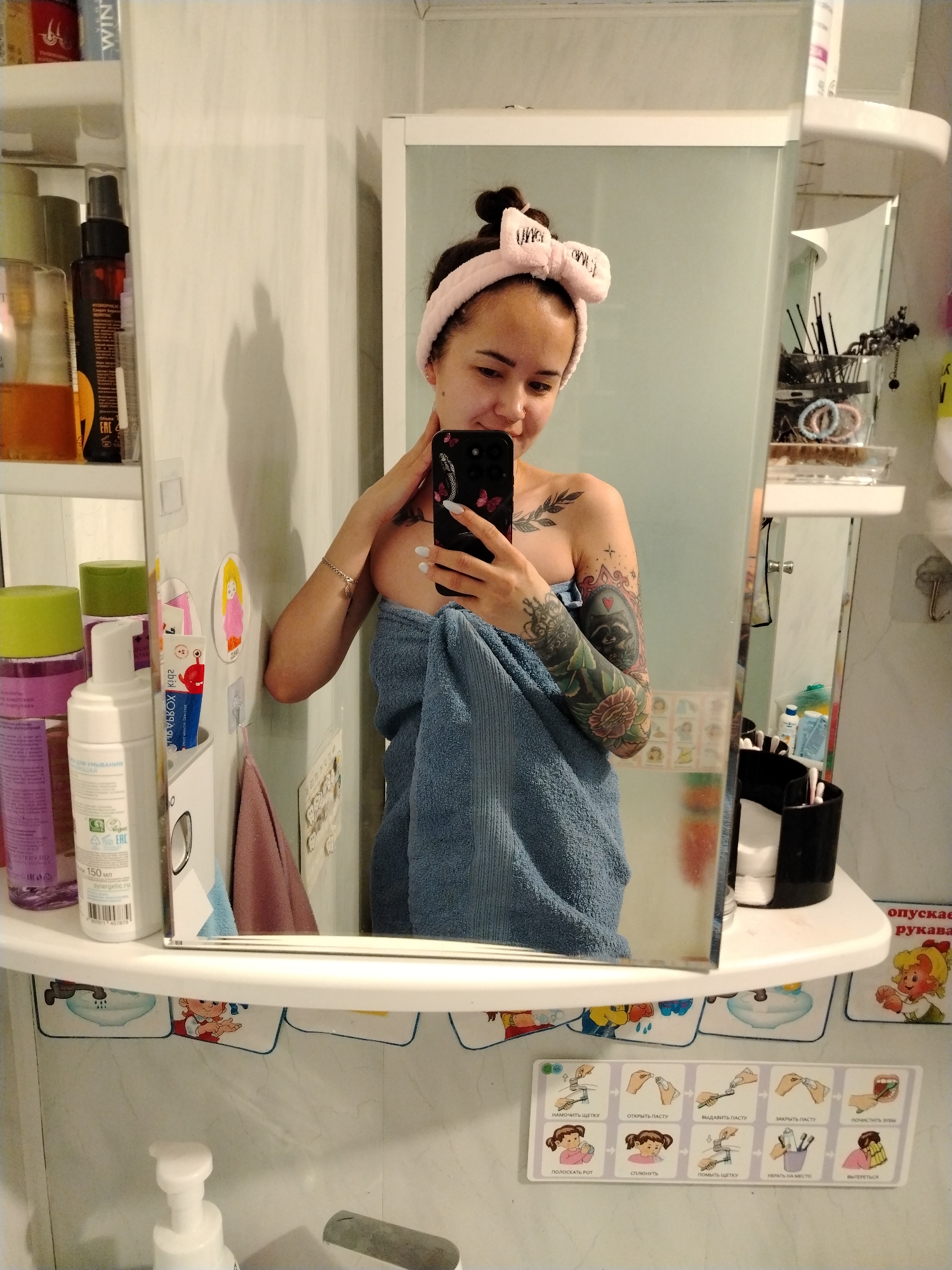 A young woman takes a mirror selfie in a bathroom in Noviy, Russia. She has dark hair tied up, wears a pink fluffy headband with a bow, and smiles softly while looking at her black smartphone, which has a butterfly-themed case. She is wrapped in a light blue towel, with visible tattoos on her arms and shoulder, including a detailed sleeve on her left arm featuring a raccoon, flowers, and an ornate frame, and a branch/leaf design on her right shoulder. A nose piercing is also visible.
The bathroom setting is clear, with white tiled walls and shelves holding various personal care products. To her left, bottles of shampoo, conditioner, and a foaming cleanser (labeled "150 мл Synergetic.ru" and "EAC") are visible, along with a product marked "WIN." To her right, reflected in the mirror, shelves hold hair accessories like scrunchies and brushes, and containers with cotton swabs. Small decorative stickers, including one of a cartoon girl, adorn the wall. A small towel hangs from a hook.
Below the main shelf, a colorful pictorial guide is affixed to the wall, illustrating hygiene steps. Visible Russian text on this guide includes: "НАМОЧИТЬ ЩЕТКУ" (Wet brush), "ОТКРЫТЬ ПАСТУ" (Open paste), "ВЫДАВИТЬ ПАСТУ" (Squeeze paste), "ЗАКРЫТЬ ПАСТУ" (Close paste), "ПОЧИСТИТЬ ЗУБЫ" (Brush teeth), "ПОЛОСКАТЬ РОТ" (Rinse mouth), "СПЛЮНУТЬ" (Spit), "ПОМЫТЬ ЩЕТКУ" (Wash brush), "УБРАТЬ НА МЕСТО" (Put away), and "ВЫТЕРЕТЬСЯ" (Wipe oneself). Partial text "опускает рукава" (lowers sleeves) is also visible on the right side of the frame.