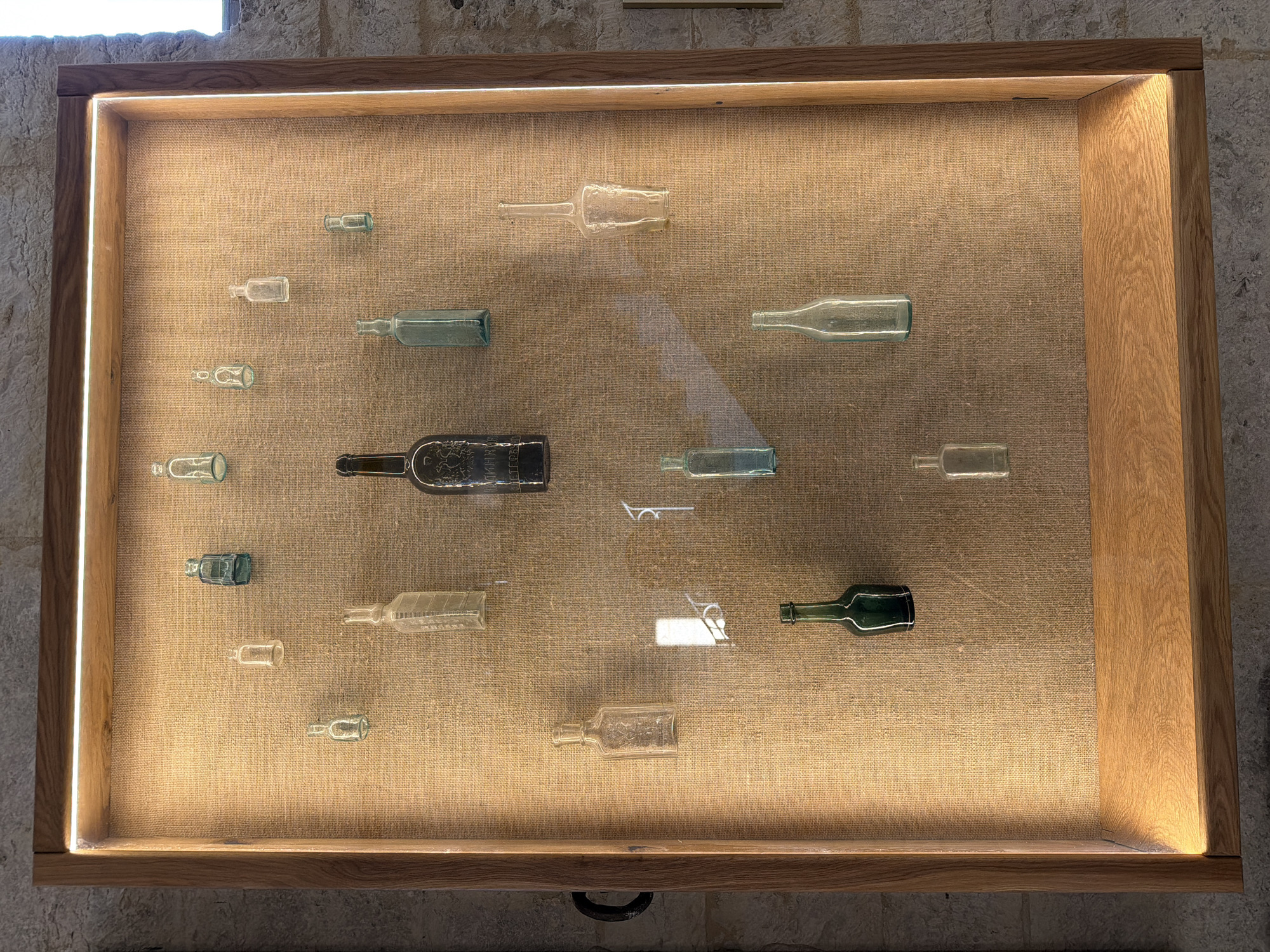 The image shows a display case containing an assortment of glass bottles of various shapes, sizes, and colors, arranged on a burlap-covered backing. The display case has a wooden frame and is illuminated by LED lights. The bottles appear to be antique, with some clear, some green, and some blue glass. They are positioned at different heights, creating a visually interesting arrangement. The background wall appears to be made of stone. It is not possible to determine the location in the city of Скорняково, Russia from the image.