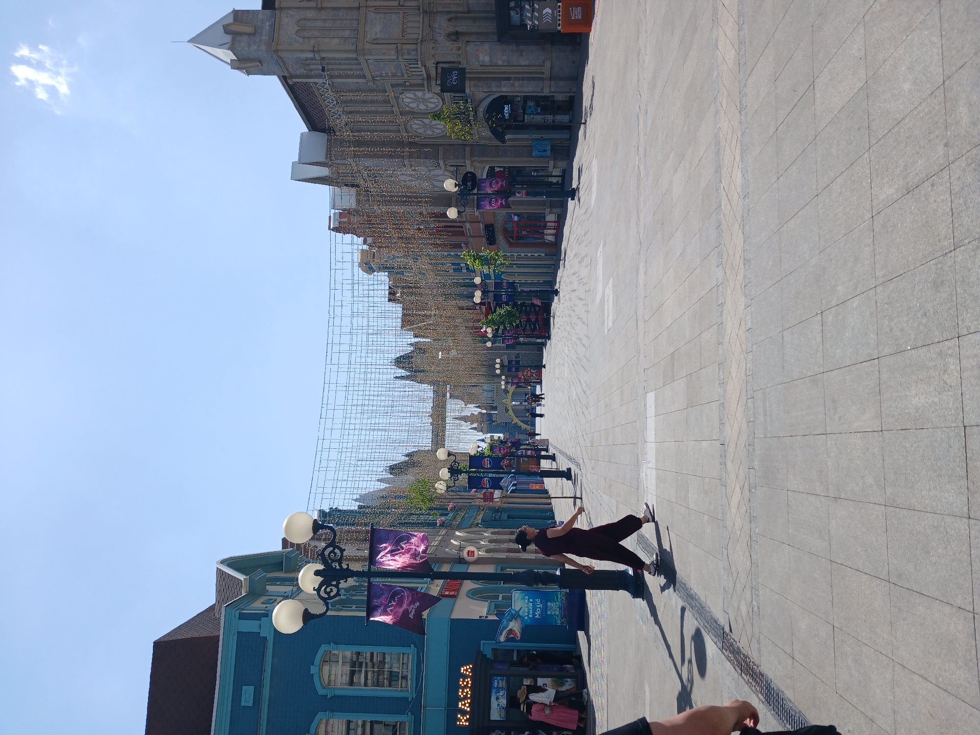 A person is captured in a dynamic pose on a sunlit, paved street lined with ornate buildings and decorative lights. The scene appears to be a pedestrian walkway or a public square, potentially in a city with European architectural influences, suggested by the facades and street lamps. The person, dressed in dark clothing, is mid-movement, with one leg extended and arms outstretched, creating a sense of motion and performance. The time of day is evident from the strong shadows cast by the person and the streetlights, indicating bright sunlight, likely midday. The sky is a clear, pale blue, suggesting fair weather.
The background features a row of buildings with intricate detailing, possibly facades of shops or entertainment venues, adorned with strings of golden lights that stretch across the street, hinting at a festive atmosphere or an event. Lampposts with multiple globes stand along the walkway. Banners are visible on some of the lampposts, displaying colorful graphics, though the text on them is not fully legible. One banner shows a graphic of what appears to be a jellyfish. Another building on the left is painted blue with white trim and features arched windows.
The paving stones of the walkway are laid in a geometric pattern, with a distinct line of darker stones marking a central division. In the foreground, a hand with a dark sleeve is visible, suggesting the perspective of someone holding the camera. The overall impression is one of a lively urban environment on a clear, sunny day, with the street serving as a stage for the person's expressive posture. The location is identified as Tashkent, Uzbekistan.