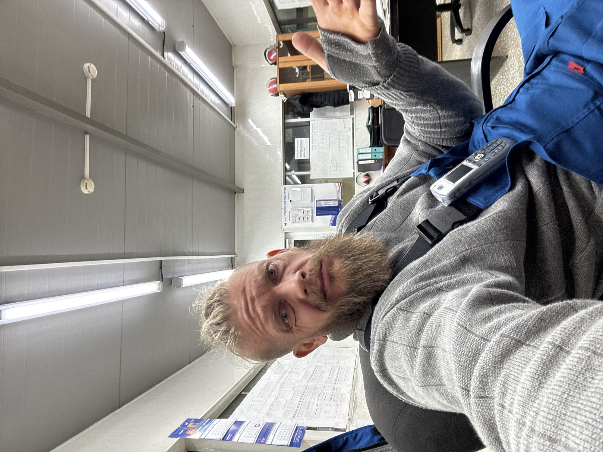 A man with blond hair and a beard wearing a grey sweater and blue work overalls is sitting in an office. He has a portable radio attached to his overalls. The office has fluorescent lighting and a white wall with a calendar. The location is Unknown, Unknown. The man is holding his hand up in the air.