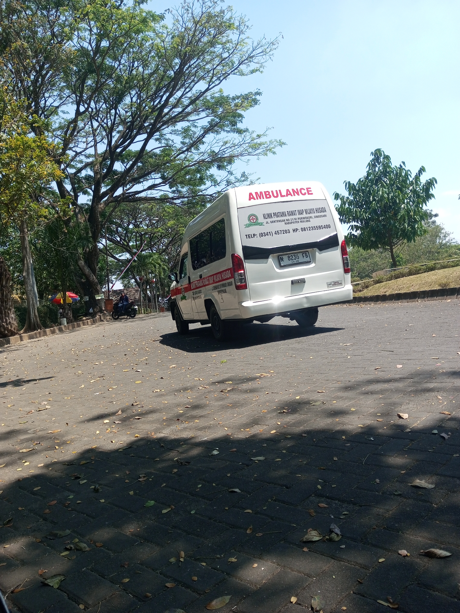 An external, eye-level, medium shot shows the rear and side of a white ambulance parked on a paved surface that is partially covered with fallen leaves. The ambulance is oriented at an angle, with its rear facing the viewer and slightly to the right. A license plate with the characters "N 8236 FB" is visible on the back of the vehicle. Text is present on the rear door of the ambulance, indicating it is an "AMBULANCE" and listing clinic information and contact details. The clinic's name is "KLINIK PRATAMA RAWAT INAP WIJAYA HUSADA," with the address "JL. Gentengan NO. 21, VI PURWOSARI, SINGOSARI, KABUPATEN MALANG." The phone number is "(0341) 457203" and the mobile number is "081235595400."
The background features a clear blue sky and large trees with lush green foliage, suggesting a sunny day. To the left of the ambulance, a red and yellow umbrella is visible, and a person on a motorcycle is seen in the mid-ground. The paved surface appears to be a pathway or road, with shadows from the trees cast across it. The environment seems to be an outdoor area, possibly a park or a facility with natural surroundings, consistent with a location in Klampok, Indonesia.
