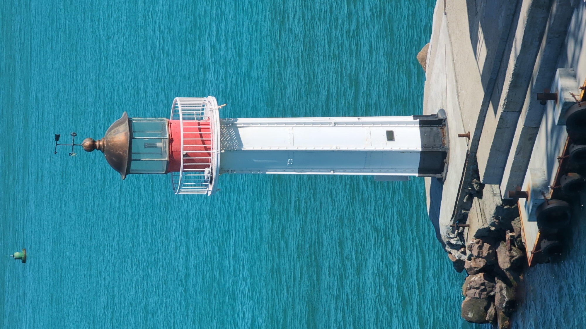 This image shows a lighthouse on a sunny day, situated on a pier extending into the turquoise waters of the Black Sea. The lighthouse is white with black and red accents and has a copper-colored lantern room. A weather vane is visible on top. The pier is made of concrete and is surrounded by rocks on one side and the sea on the other. In the distance, a green buoy is visible in the water. The scene appears to be the approach to the port of Burgas, Bulgaria.