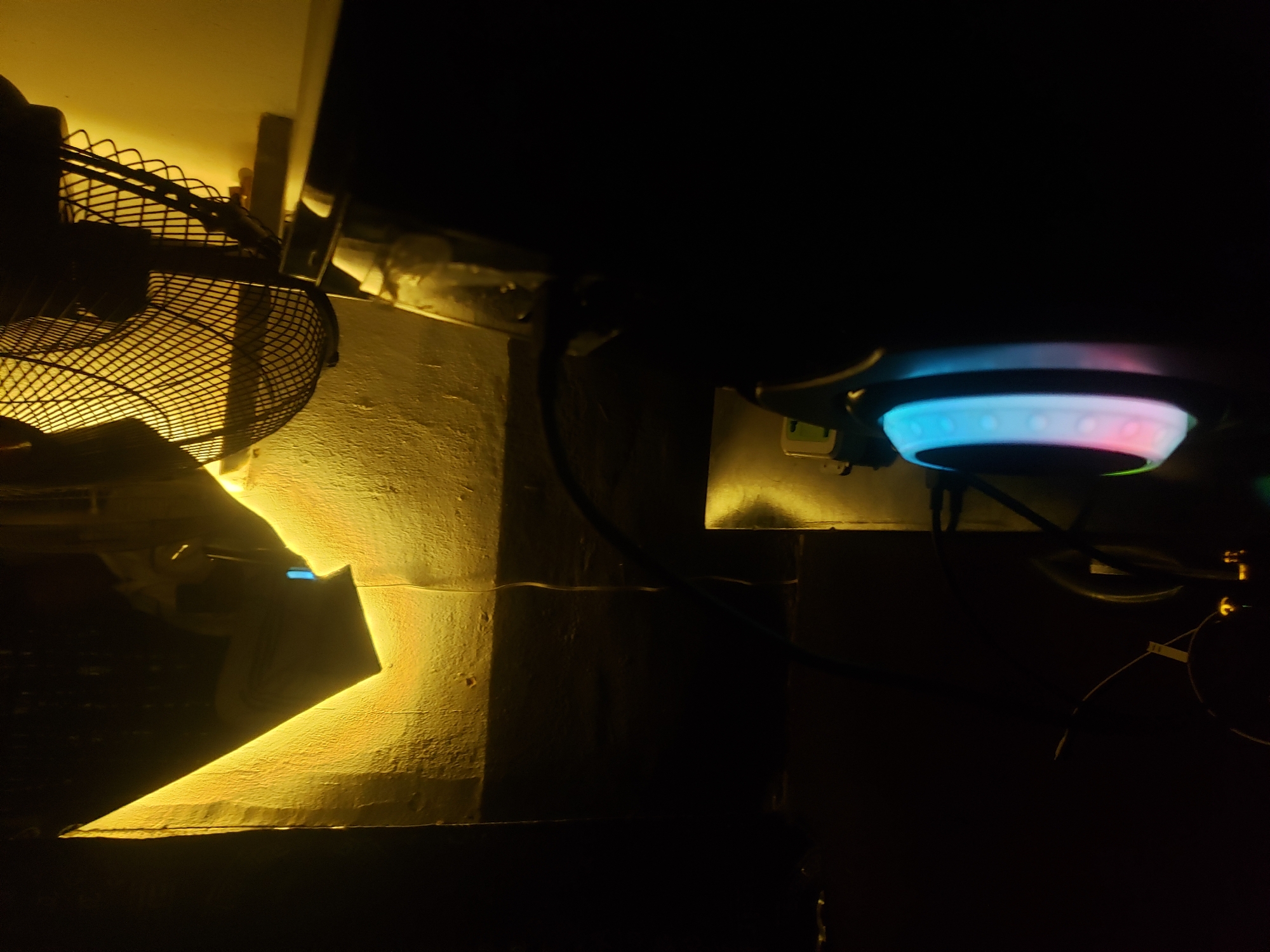 In an indoor, dimly lit environment, a close-up shot focuses on various objects. To the left, a textured wall is illuminated by a bright yellow light source, creating a sharp, angular shadow. A portion of a fan with a dark grill is visible at the top right, casting shadows. In the foreground, a pair of headphones with colorful LED lights is the most prominent object, with a blue and pink gradient visible on the earcups. Cables are also present, snaking across the scene. The overall scene is dark with selective illumination, suggesting a domestic or gaming setup.