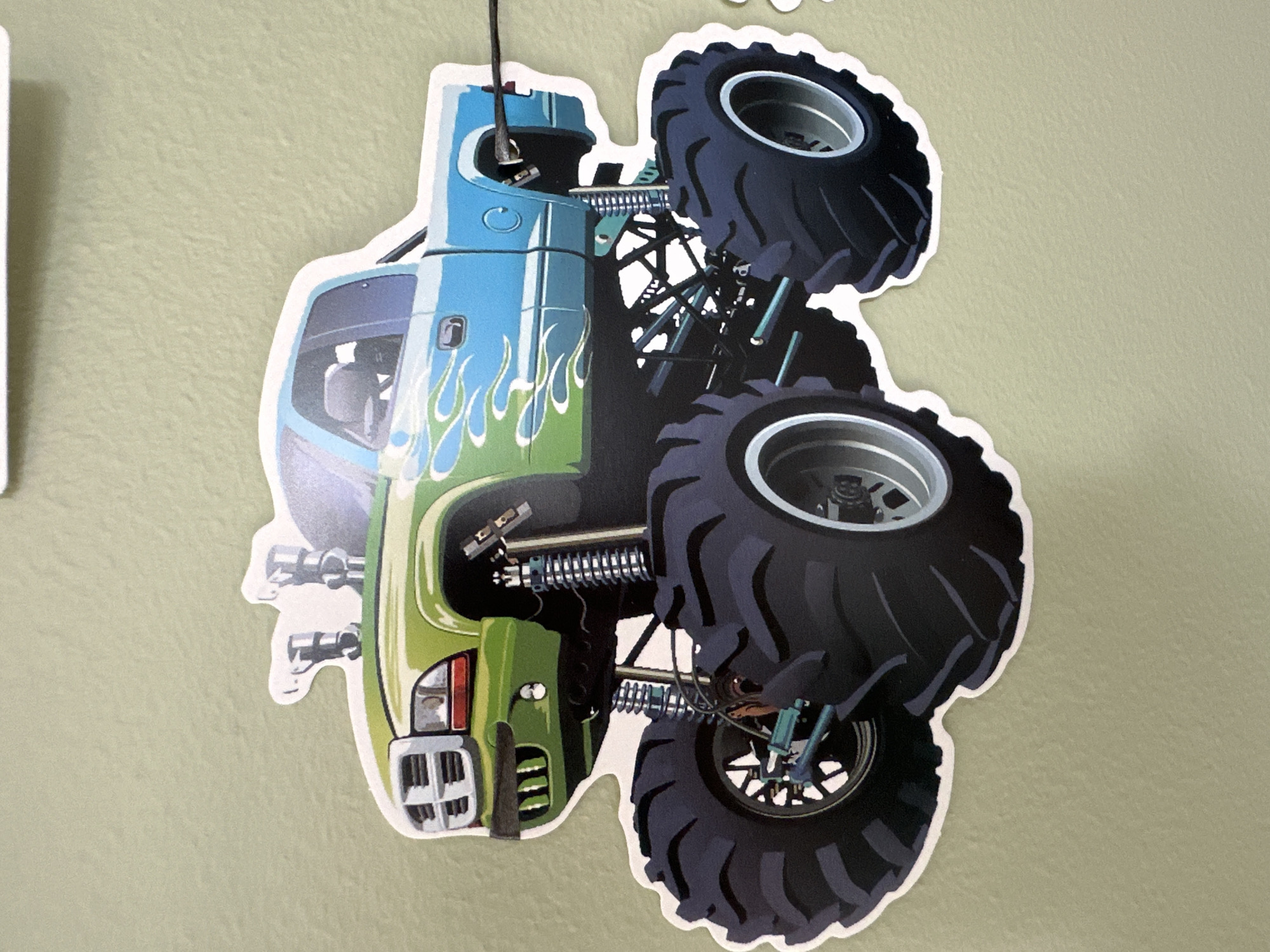 This media file is a close-up, high-angle shot of a sticker of a monster truck. The truck is depicted as being mid-air, with its front end angled upwards. The truck has a green and blue color scheme, with stylized flames adorning the side. It features large, black, knobby tires and exposed suspension components, indicative of its off-road capabilities.
The sticker is attached to a pale green wall, which appears to have a slightly textured surface. A thin black string hangs from the top of the sticker, suggesting it may be part of a mobile or decoration.
The lighting appears to be even and diffused, with no harsh shadows, implying an indoor setting or an overcast day. There is no visible text in the image. The overall impression is of a colorful, dynamic toy or decoration.