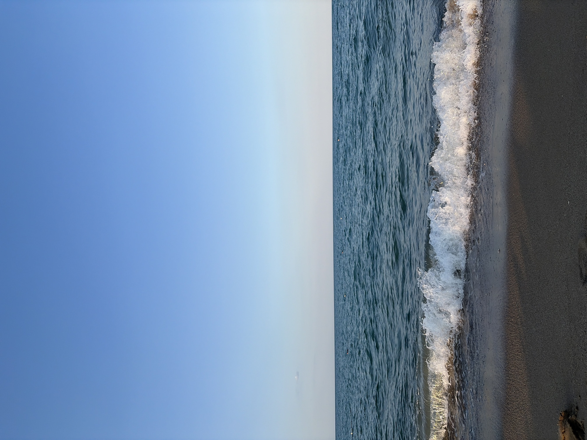The image depicts a tranquil scene of the Black Sea coast in Obzor, Bulgaria. The horizon line is clearly defined, separating the vast expanse of a clear, pale blue sky from the deep blue waters of the sea. A gentle wave is breaking on a sandy beach in the foreground, its white foam contrasting with the dark, wet sand. The water appears calm, with subtle ripples and a few distant buoys visible on the surface. The overall impression is one of a peaceful, natural setting, characteristic of the Obzor coastline.