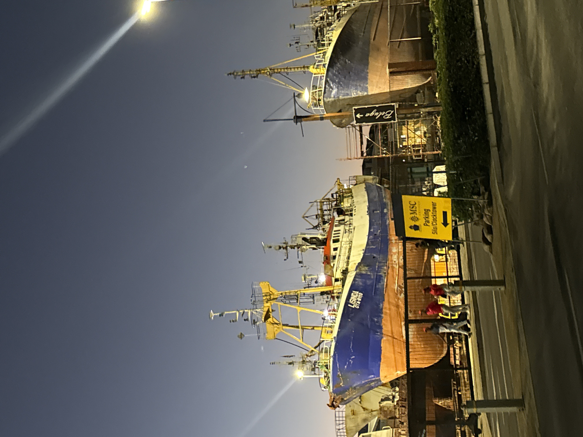 The image depicts a harbor setting in Cape Town, South Africa at dusk. Several large fishing vessels are visible, likely undergoing maintenance or repair. One vessel, named "LISINDA GTA-6110" with a blue and orange hull, is prominently featured. A yellow sign indicates directions for "MSC Parking Silo/Clocktower." People are present behind a fence, likely workers. The scene is illuminated by artificial lighting, including a street lamp and lights on the vessels themselves. Scaffolding surrounds the vessels, supporting the maintenance activities. Signage for "Beluga" is also visible. The environment is urban and industrial.