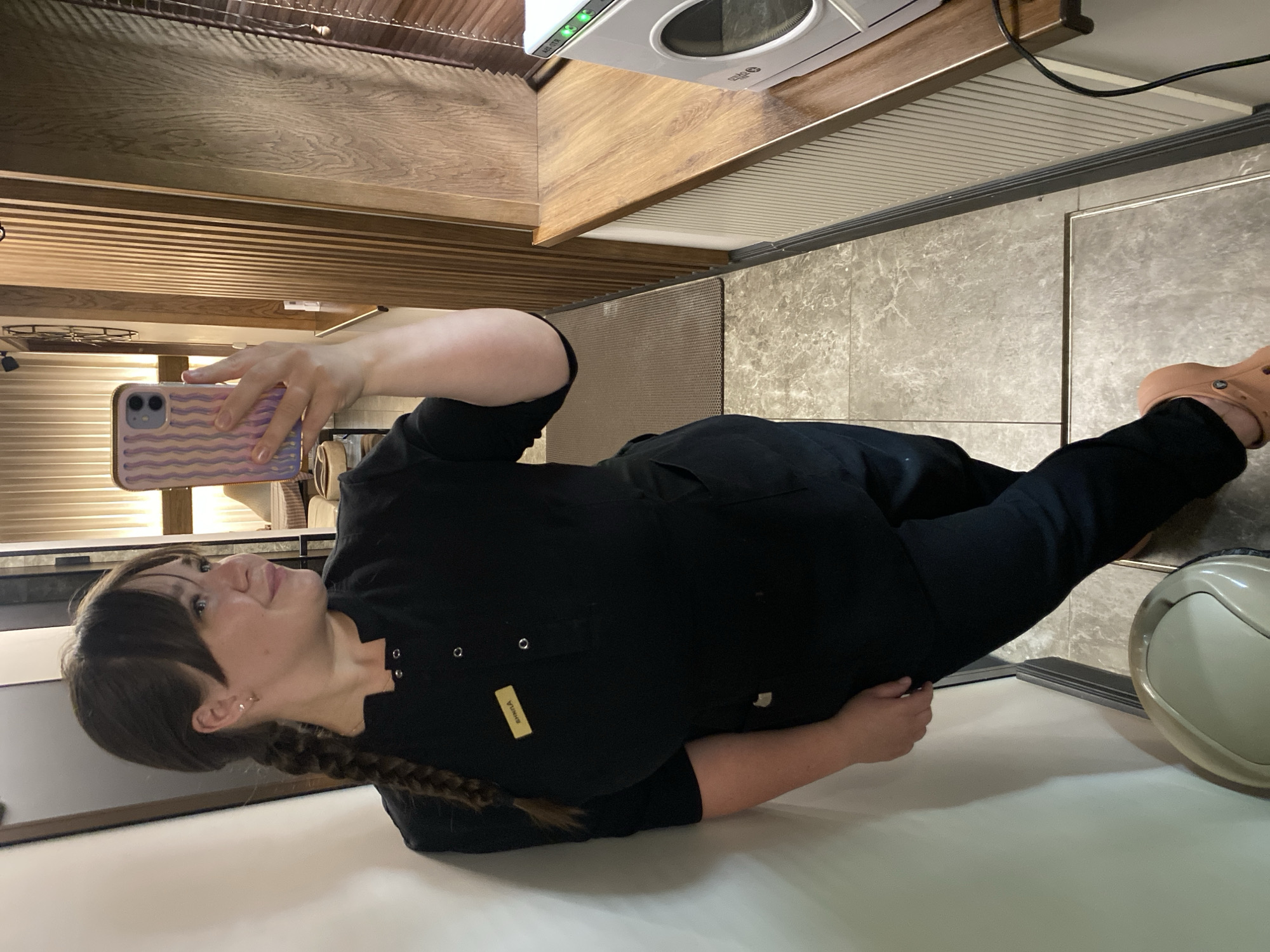 A woman, identified by a name tag as "SHHRILA", is standing in a hallway, taking a selfie with her phone. She is wearing a black uniform, consisting of a collared shirt with buttons and matching pants. Her hair is dark brown and styled in a braid. She is leaning against a white wall, with her left leg bent and her right leg extended, wearing tan Crocs. The hallway has marble-patterned flooring and a textured rug. To the right, there is a wooden wall feature and a wooden shelf holding a white appliance with a circular window, possibly a sterilizer. The overall environment suggests a commercial establishment, such as a spa or salon, possibly located in Votkinsk, Russia.