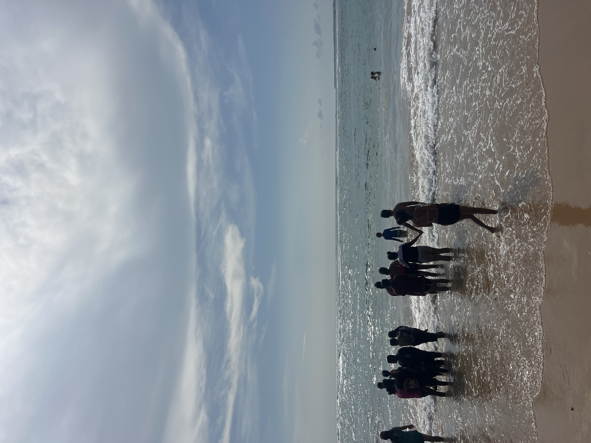 In Hikkaduwa, Sri Lanka, a group of individuals is gathered on a beach, some standing in the shallow ocean water with gentle waves lapping at their ankles. The sky above is partly cloudy, with a blend of blue sky and wispy white clouds. The sun reflects brightly off the ocean's surface, creating a shimmering effect. Several people are visible in the background, some swimming further out in the sea. The sand on the beach appears wet and dark near the water's edge, transitioning to a lighter, drier hue further up. The scene captures a moment of leisure and enjoyment at the seaside.