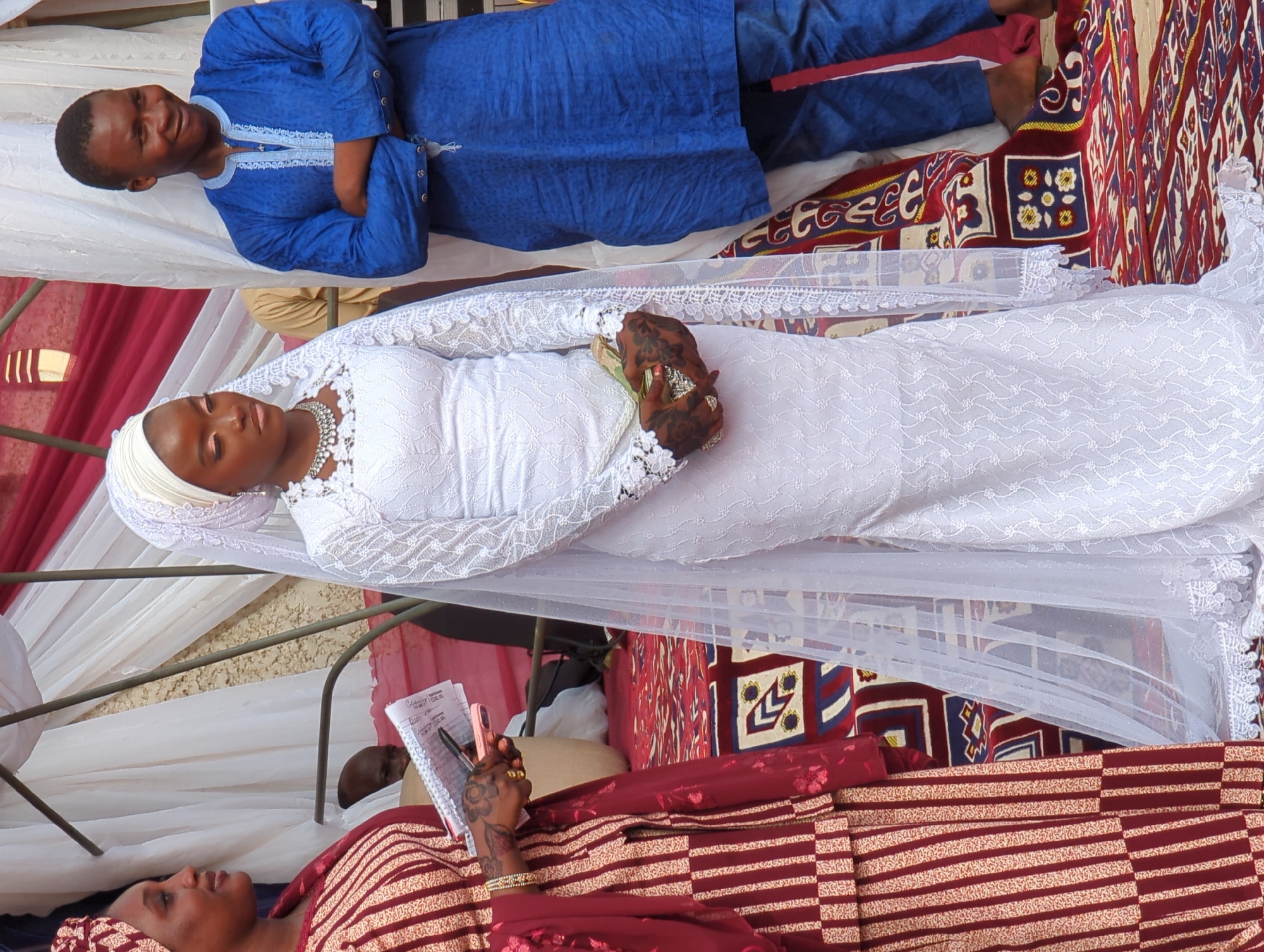 The image depicts an outdoor or semi-outdoor event in Kabala, Nigeria, with three main individuals positioned on an ornate red and blue patterned rug.
On the left, an adult female with dark skin and a slight smile looks towards the left. She wears a long dress with vertical maroon and beige stripes, a maroon headwrap, and a matching maroon shawl draped over her left arm. Her left hand, adorned with intricate dark henna patterns, holds a pink smartphone and rests on an open spiral-bound notebook containing visible text. A gold bracelet and a gold ring are visible on her left hand.
In the center stands a young adult female, appearing to be a bride, with dark skin and a focused expression. She wears a fitted, floor-length white lace wedding gown with long sleeves, a sheer white veil with lace trim draped over a white headwrap, and a prominent silver choker necklace. Her hands, also adorned with dark, intricate patterns, are clasped together at waist height, holding a small, silver-embellished clutch purse, from which several green banknotes are partially visible.
On the right, an adult male with dark skin and a broad smile looks towards the right. He wears a royal blue long-sleeved tunic with light blue embroidery around the collar and chest, paired with dark blue trousers. His arms are crossed over his chest, and his bare feet or dark sandals are visible on the rug.
The background features white and maroon draped fabrics, indicating a decorated event space or canopy structure, supported by visible metal poles. A bald head of a person is partially visible behind the left woman, and a glimpse of a person in a tan shirt is seen behind the bride.