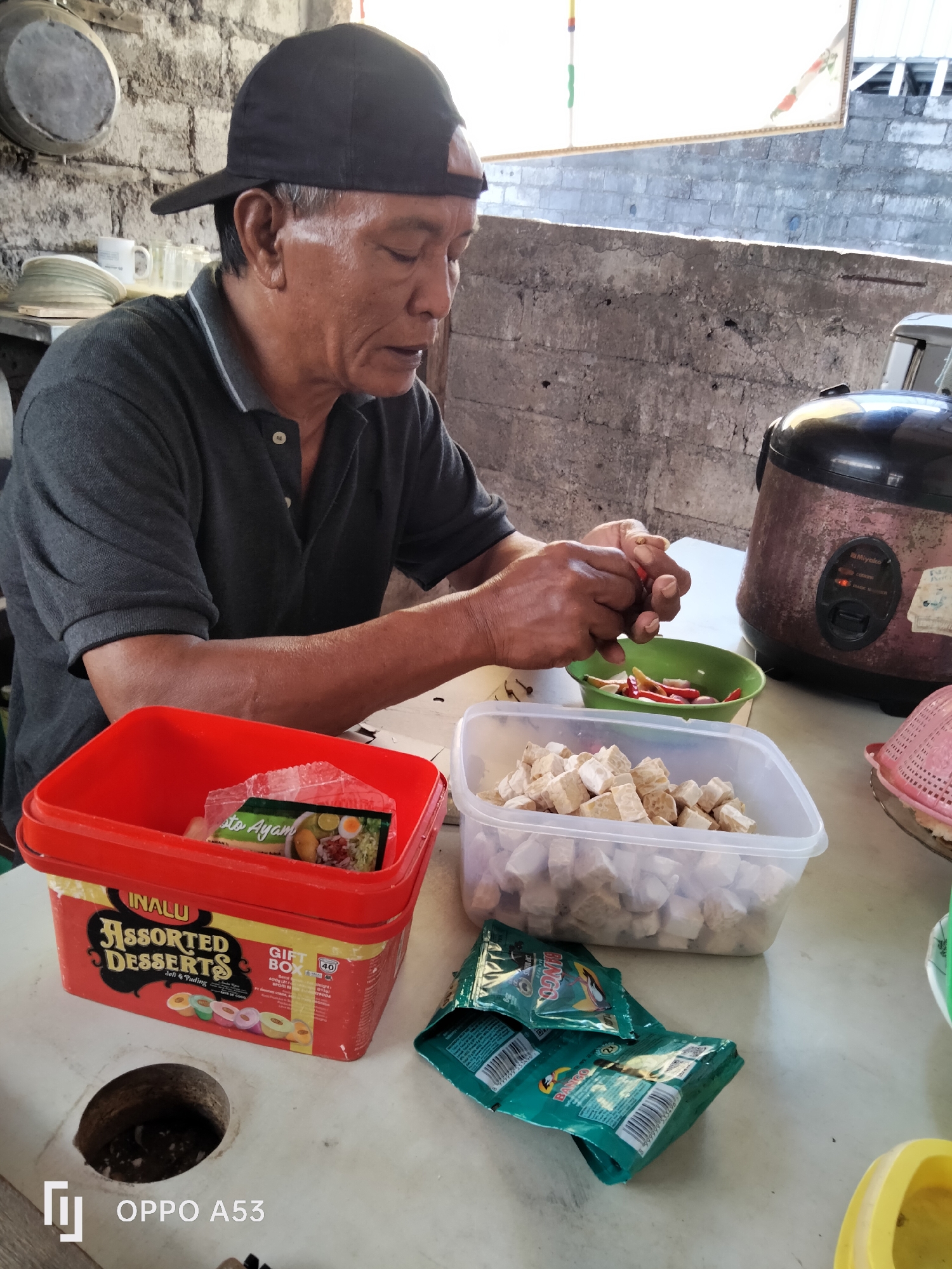 A man wearing a dark grey polo shirt and a black baseball cap is seated at a white table, preparing food. His eyes are closed as he holds red chilies in his hands, seemingly in the process of chopping them. In front of him, a green bowl contains sliced red chilies and onions. To his right, a clear plastic container holds cubed tempeh. A red plastic tub labeled "INALU Assorted Desserts" is to his left, with a small packet of "Soto Ayam" inside. Behind the man, a large, stained rice cooker with a "Miyako" logo is visible. The background consists of a rough, grey concrete wall and a partial view of the sky. The table surface is cluttered with various food items and containers, suggesting a casual outdoor cooking or meal preparation setting. The scene is illuminated by natural daylight. The location appears to be an outdoor or semi-outdoor area with a rustic, functional aesthetic.