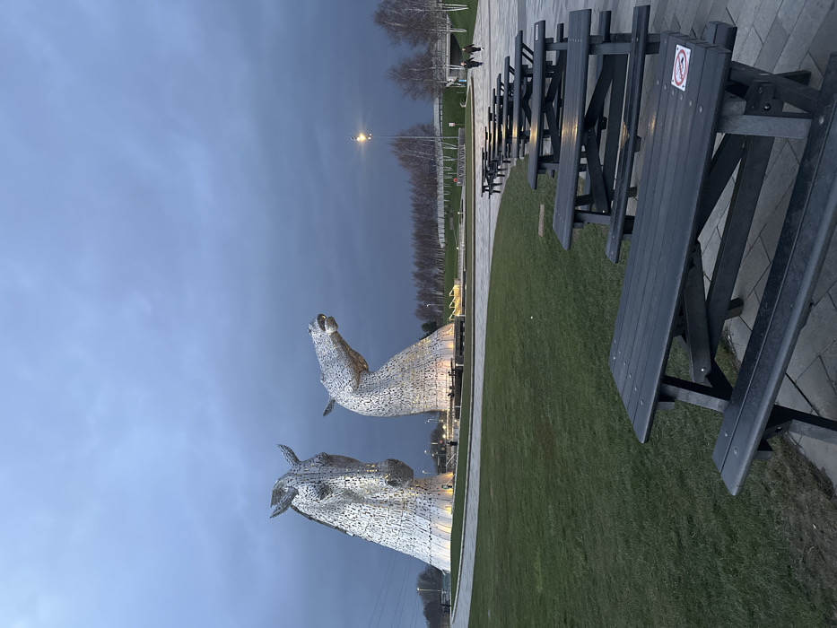 The image depicts an outdoor scene at The Helix Park in Falkirk, United Kingdom, featuring The Kelpies sculptures at dusk or dawn under an overcast, dark grey sky. In the mid-ground, two massive, stylized horse head sculptures, constructed from reflective metallic panels, stand illuminated from below. The left sculpture faces generally forward, while the right sculpture's head is angled upwards and to the right. Both sculptures are situated on circular bases surrounded by paved areas and manicured green grass.
In the foreground, a row of approximately eight dark-colored, unoccupied wooden picnic tables with attached benches extends from the lower right towards the mid-ground, set on a grey paved surface. One table in the immediate foreground displays a white sticker with a red prohibition symbol. The ground transitions to a well-maintained green grass lawn to the left.
In the background, beyond the sculptures, a paved pathway is visible. Four human figures are present: two indistinct individuals near the base of the left sculpture, and two individuals walking away from the camera along the pathway in the far right mid-ground. A tall streetlamp with a bright light is visible to the right of the sculptures. A line of bare deciduous trees runs horizontally across the distant background, with power lines and other urban infrastructure faintly visible on the far left. The overall lighting is low, emphasizing the illuminated sculptures against the dark, featureless sky.
