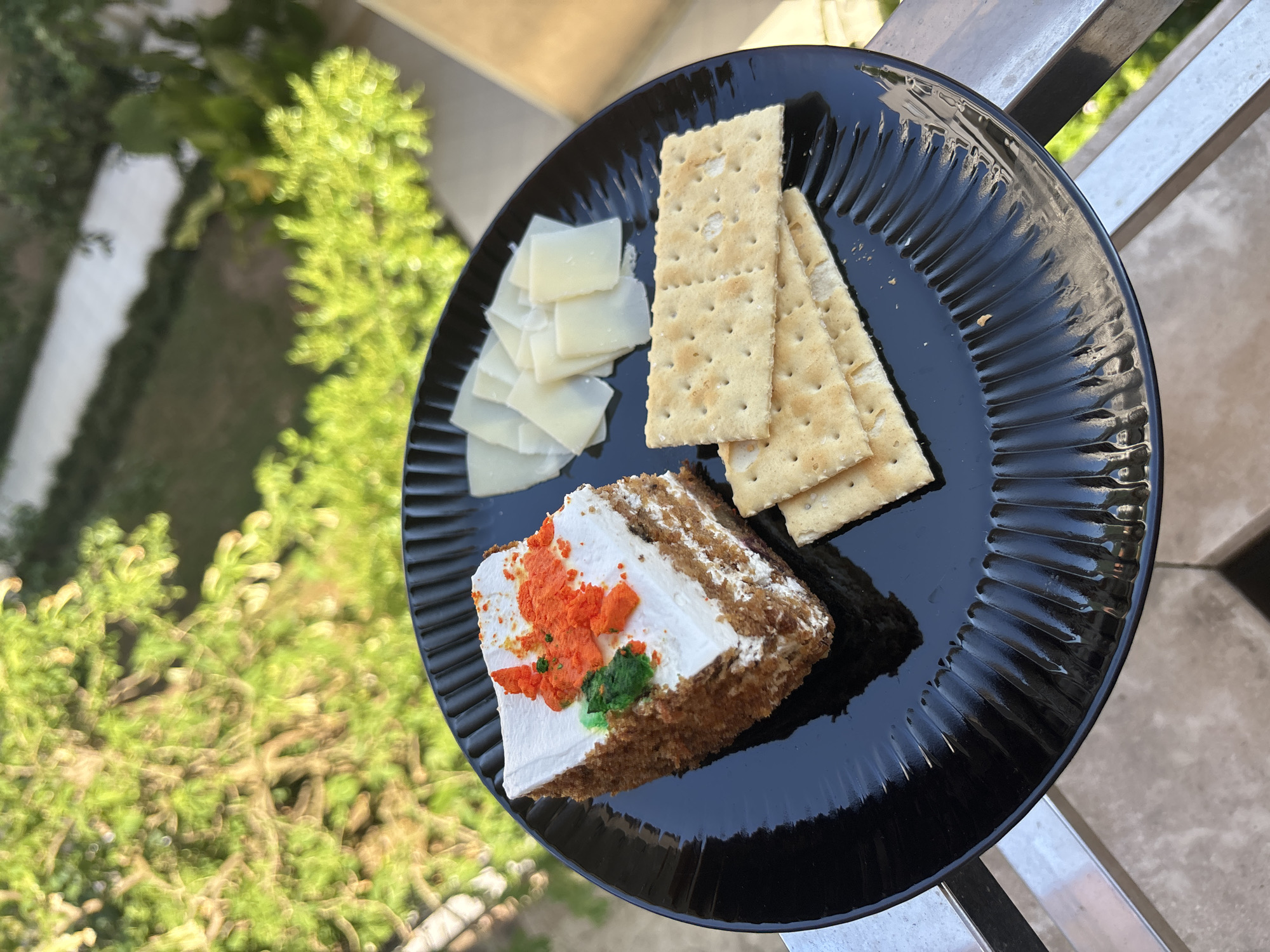 A slice of carrot cake topped with white frosting and orange and green sprinkles is served on a black ribbed plate. Next to the cake are several thin slices of cheese and three square crackers. The plate is on a metal railing outdoors, with green foliage and a body of water visible in the background. This location appears to be a balcony or patio in Budva, Montenegro.