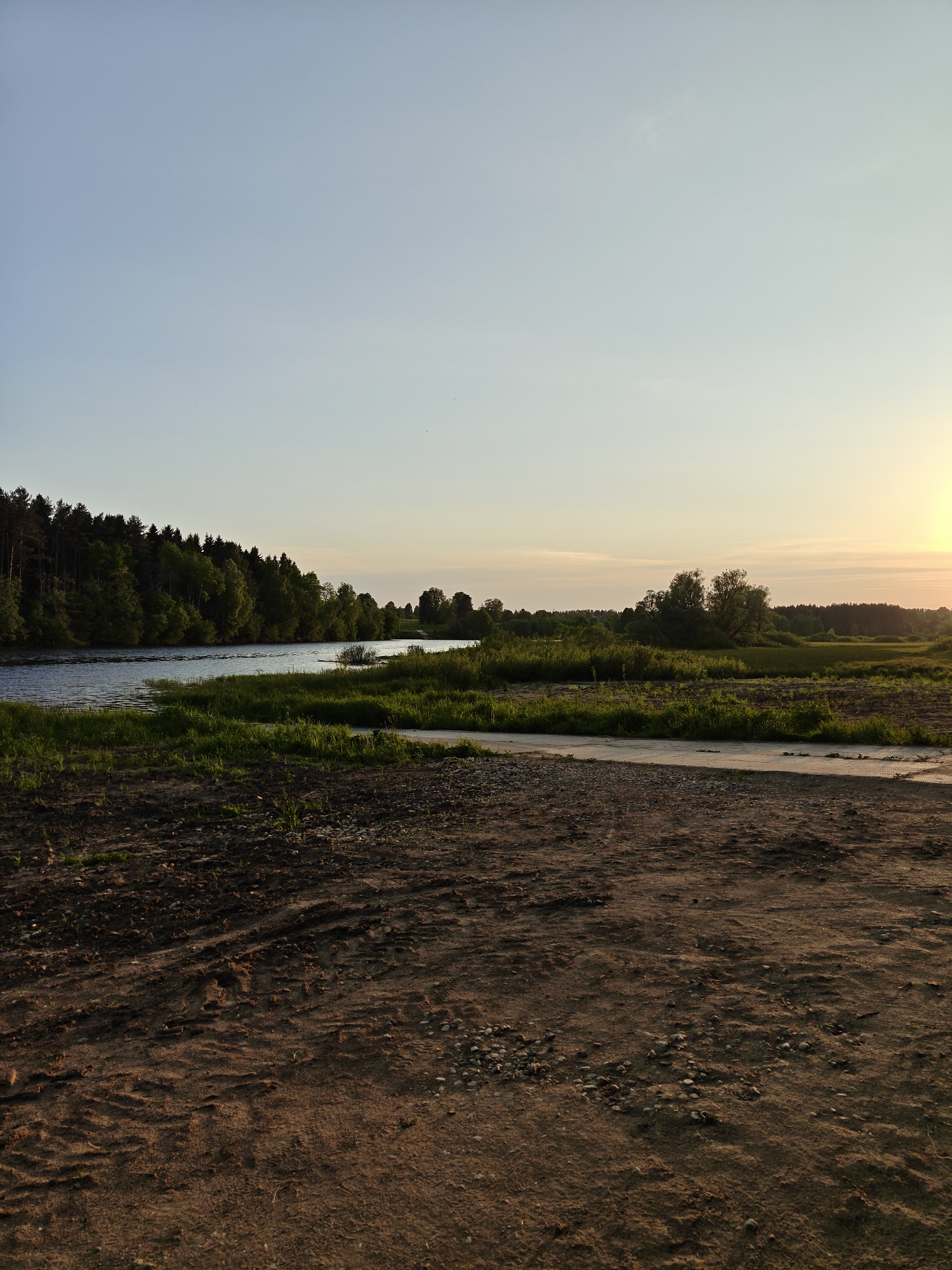 The image presents a landscape scene in Мышкино, Russia, captured during sunset. The foreground features a dirt road with visible tire tracks and scattered pebbles. Beyond the road, there's a grassy area leading to a body of water, possibly a river or a wide stream. The water reflects the sky, and the opposite bank is lined with dense trees. The sky transitions from light blue to a warmer tone as the sun sets on the right side of the frame. No people or animals are visible in the image.