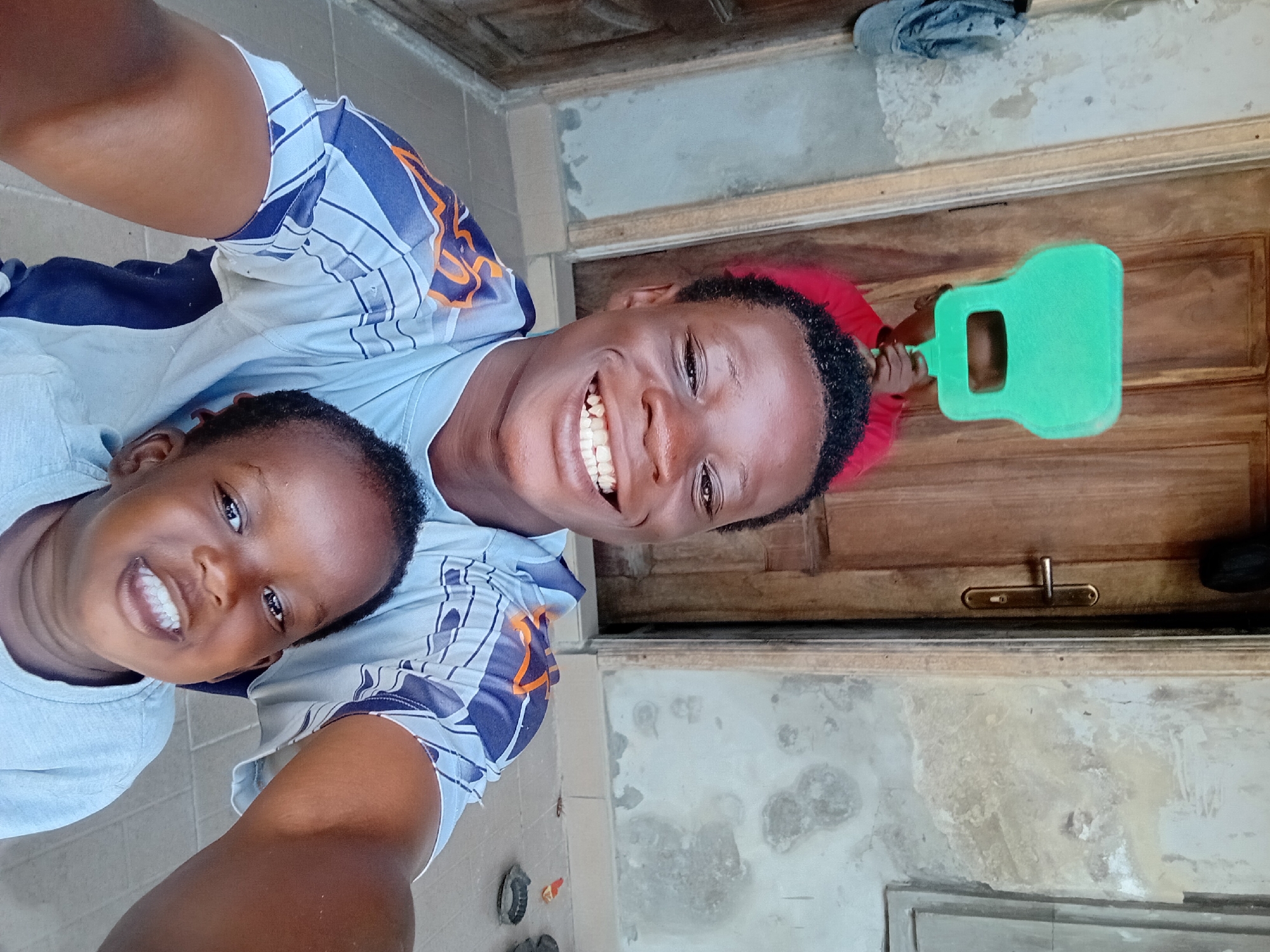 Two young Black males are posing for a selfie outdoors. The male in the foreground is on the left and is smiling broadly at the camera. He is wearing a light blue and white striped t-shirt with blue and orange accents. To his right and slightly behind him is a younger male, also smiling. The younger male is wearing a light gray t-shirt. Behind the two males, a third individual is partially visible wearing a bright red shirt and holding a green plastic item, possibly a toy or prop, in front of their face. The background consists of a wooden door with a brass handle, weathered walls, and a tiled floor. A cap hangs on a hook to the left of the door. The overall scene appears to be a casual, outdoor setting, likely in a residential area.