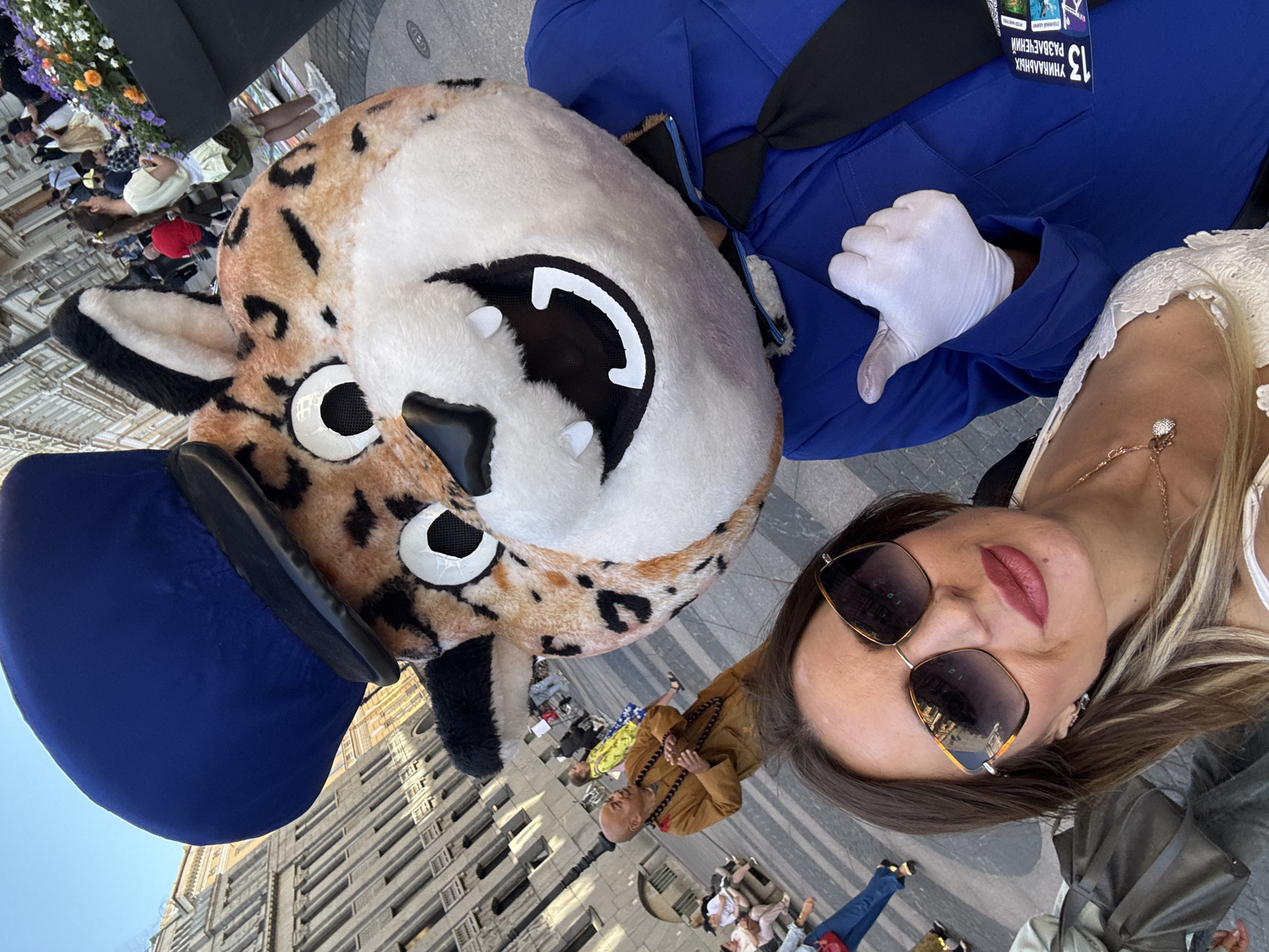The image captures a selfie taken by a woman with blonde hair, wearing large sunglasses and a white lace top. She is posing with a person in a mascot costume of a leopard wearing a blue uniform and hat. The mascot is giving a thumbs-up gesture. In the background, a man in a monk's robe is visible, along with other people walking and sitting. The scene appears to be taking place on a paved area, possibly a plaza or street, with a building in the background that resembles architecture found in Saint Petersburg, Russia. There are also flower arrangements in the foreground. The overall setting suggests an outdoor event or gathering in a city.