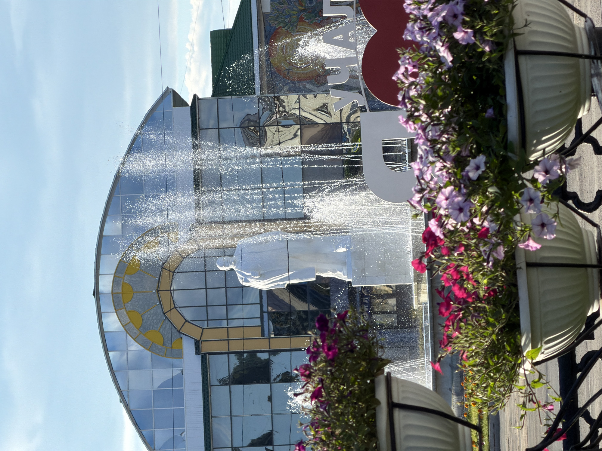 A fountain sprays water in front of a modern building with a curved glass facade. The building has a large, circular window with a yellow and gray design. On the facade, there is a large, white sculptural figure of a person. Above the figure and to the right, large white letters spell out "АРХ" and part of another word. Behind the sculpture, a colorful mosaic is visible on the building. In the foreground, several large planters are filled with blooming pink and purple flowers. The scene appears to be outdoors, with a bright sky and sunlight reflecting off the glass and water. The image is oriented vertically.
