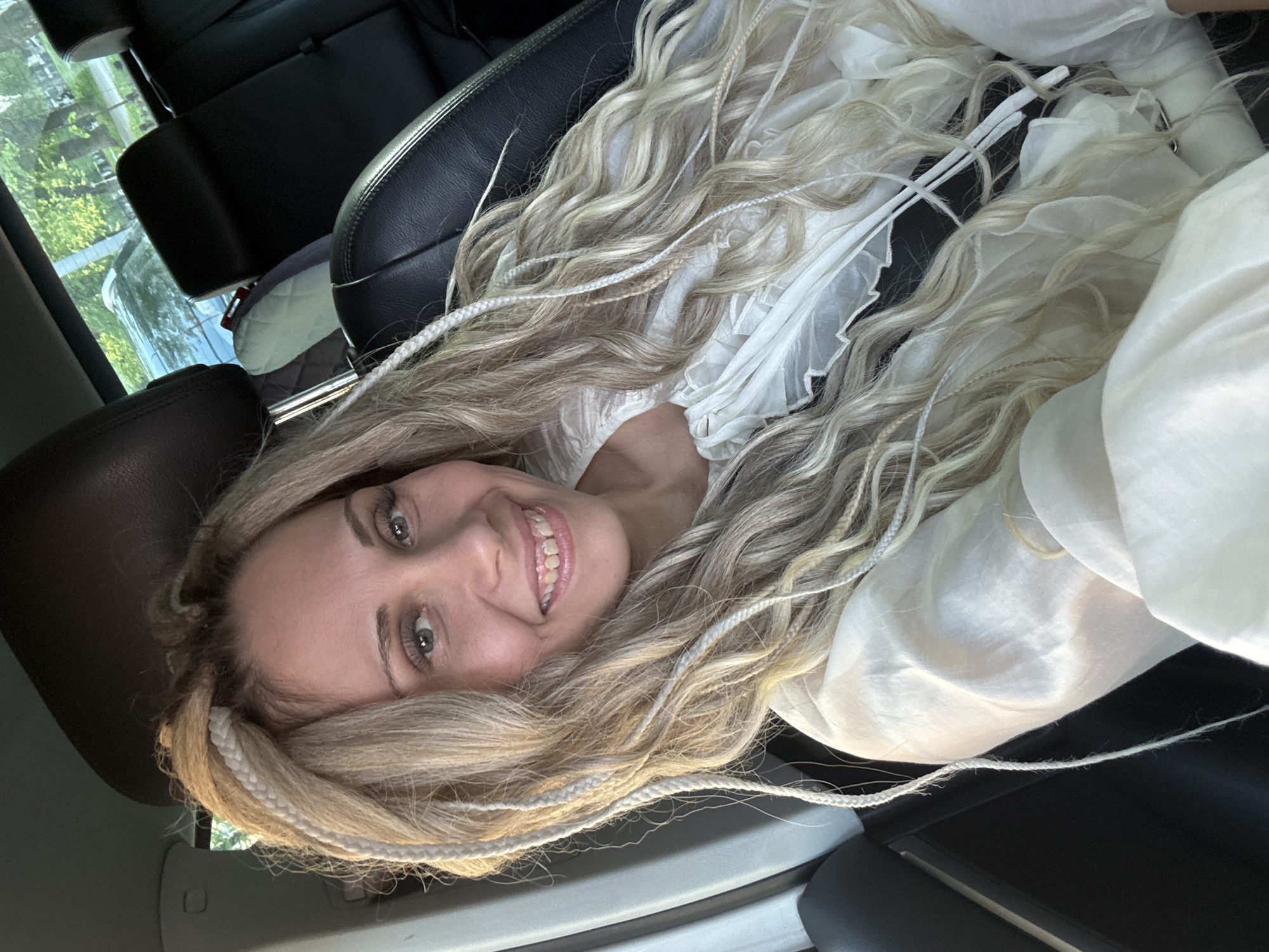 A young, fair-skinned woman with long, wavy, blonde hair interwoven with thin, white braids is reclining in the passenger seat of a vehicle. She is wearing a white blouse with ruffles. She is smiling and looking directly at the camera. The interior of the vehicle is visible, including black leather seats and a view through the window showing greenery. The location is unknown and there is no information in the image to indicate it is Rusanovo, Russia.