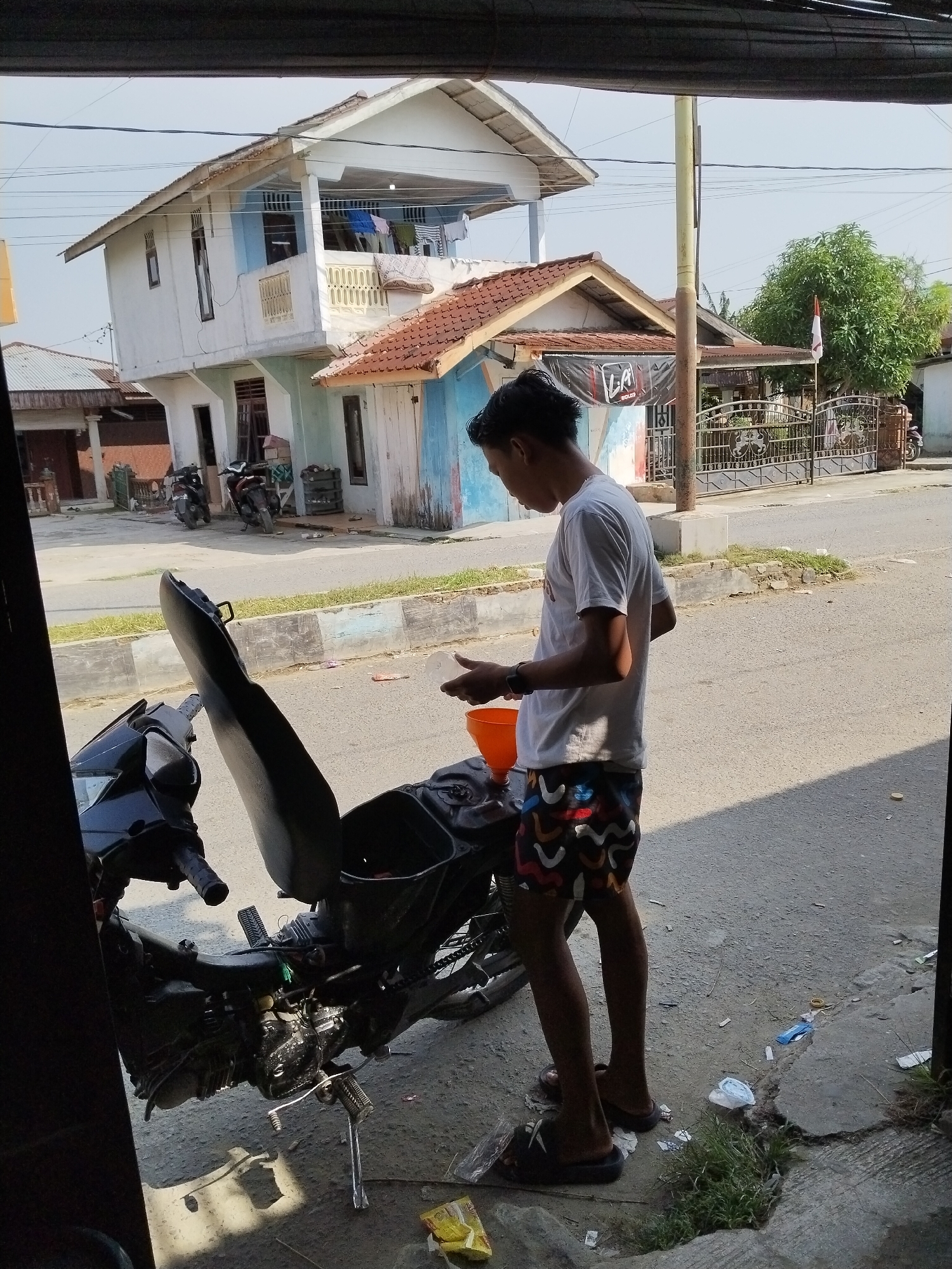 A young man stands beside a motorcycle in front of a building in Sawit Seberang, Indonesia. He is wearing a white t-shirt and patterned shorts, and is holding a white cup and an orange funnel. The motorcycle is black with its seat open, revealing the engine. In the background, there is a two-story house with laundry hanging on a balcony. The street is paved and there are some trash items on the ground. The scene appears to be in a residential area.