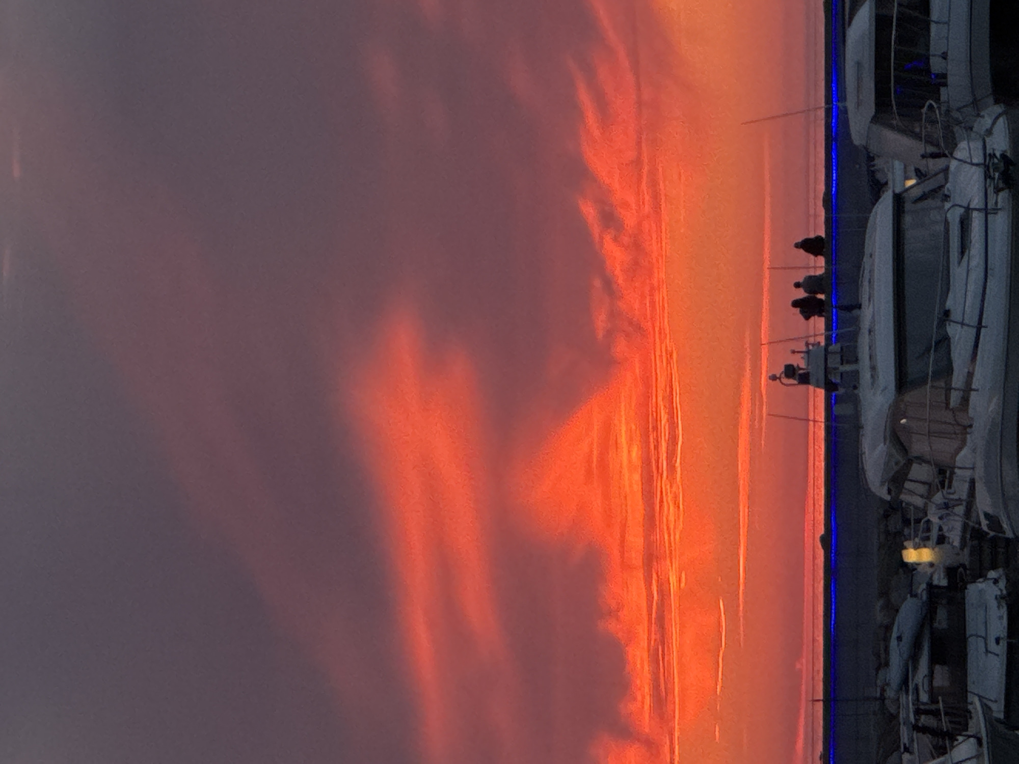This image depicts a stunning sunset or sunrise over a marina in Tallinn, Estonia. The sky, dominating the left two-thirds of the frame, is ablaze with vibrant and intense hues of orange, red, and purple. Wisps and streaks of clouds are dramatically illuminated by the low sun, creating a fiery and ethereal display.
In the lower right portion of the image, a dark pier or shoreline extends horizontally. A distinctive thin blue light runs along its edge. Several silhouetted figures of people are visible on this pier, appearing to be observing the magnificent sky. Their forms are indistinct due to the strong backlighting.
To the far right, a row of large boats or yachts is docked. These vessels are also rendered in dark silhouette, though structural details like railings and cabins are discernible. One boat near the center right emits a soft, yellowish glow from an interior or deck light. A narrow strip of dark water is visible in front of the pier.
The scene exudes a peaceful and awe-inspiring atmosphere, highlighting the dramatic beauty of dawn or dusk over a harbor. The striking contrast between the brilliantly colored sky and the shadowed foreground elements is a notable feature. No visible text is present in the image.