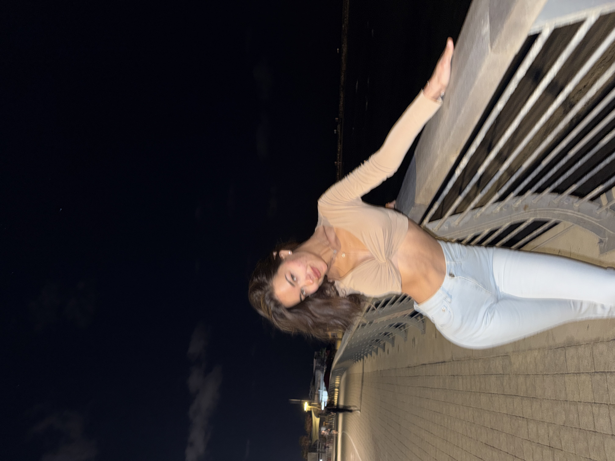 A young adult female is depicted at night on a paved waterfront promenade in an unknown city. She is positioned centrally, leaning with her left arm on a modern metal railing that separates the walkway from a dark body of water. The female has long, dark brown hair, wears a light tan long-sleeved crop top with a twisted front, and light blue high-waisted jeans. She is looking towards the camera with a slight smile, revealing small stud earrings and a delicate necklace.
To her left, the stone-paved walkway extends into the distance, illuminated by artificial streetlights, where a blurred human figure is visible walking away from the camera. To her right, beyond the railing, is a vast, unlit body of water, appearing black. The night sky above is dark, with faint cloud formations visible in the upper left. Artificial light sources illuminate the subject and the immediate foreground, creating strong contrast with the dark background elements.