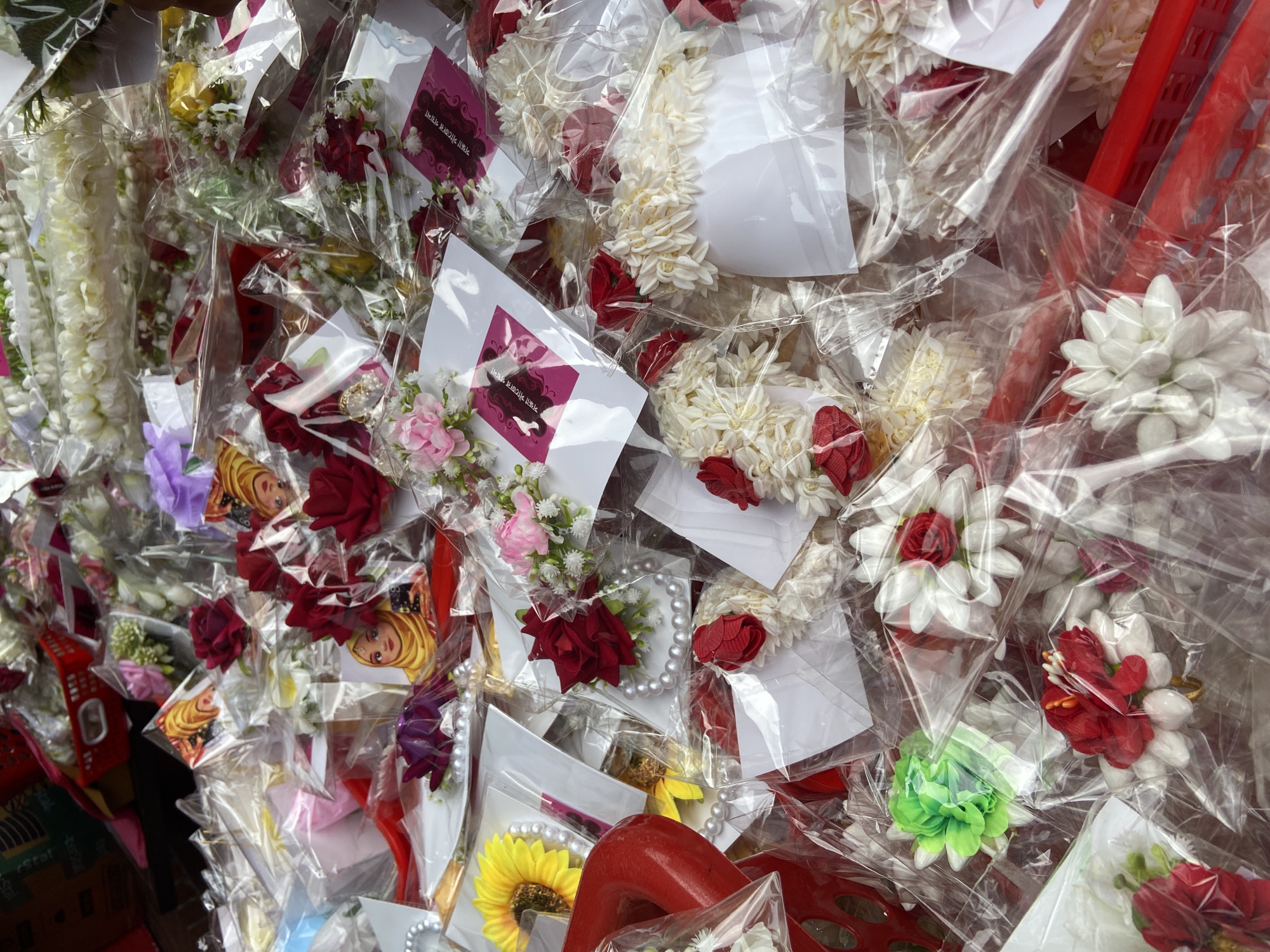 The image shows a collection of flower garlands and floral decorations, each individually packaged in clear plastic. These items appear to be for sale, possibly for religious or celebratory occasions. Many of the garlands consist of white flowers accented with red roses. Some of the packages also contain small cards with text in Bengali. The scene is likely located in Dhaka, Bangladesh, given the Bengali script and the prevalence of flower garlands in South Asian religious practices. The items are densely packed together, suggesting a retail display. Some of the arrangements include small pearls.