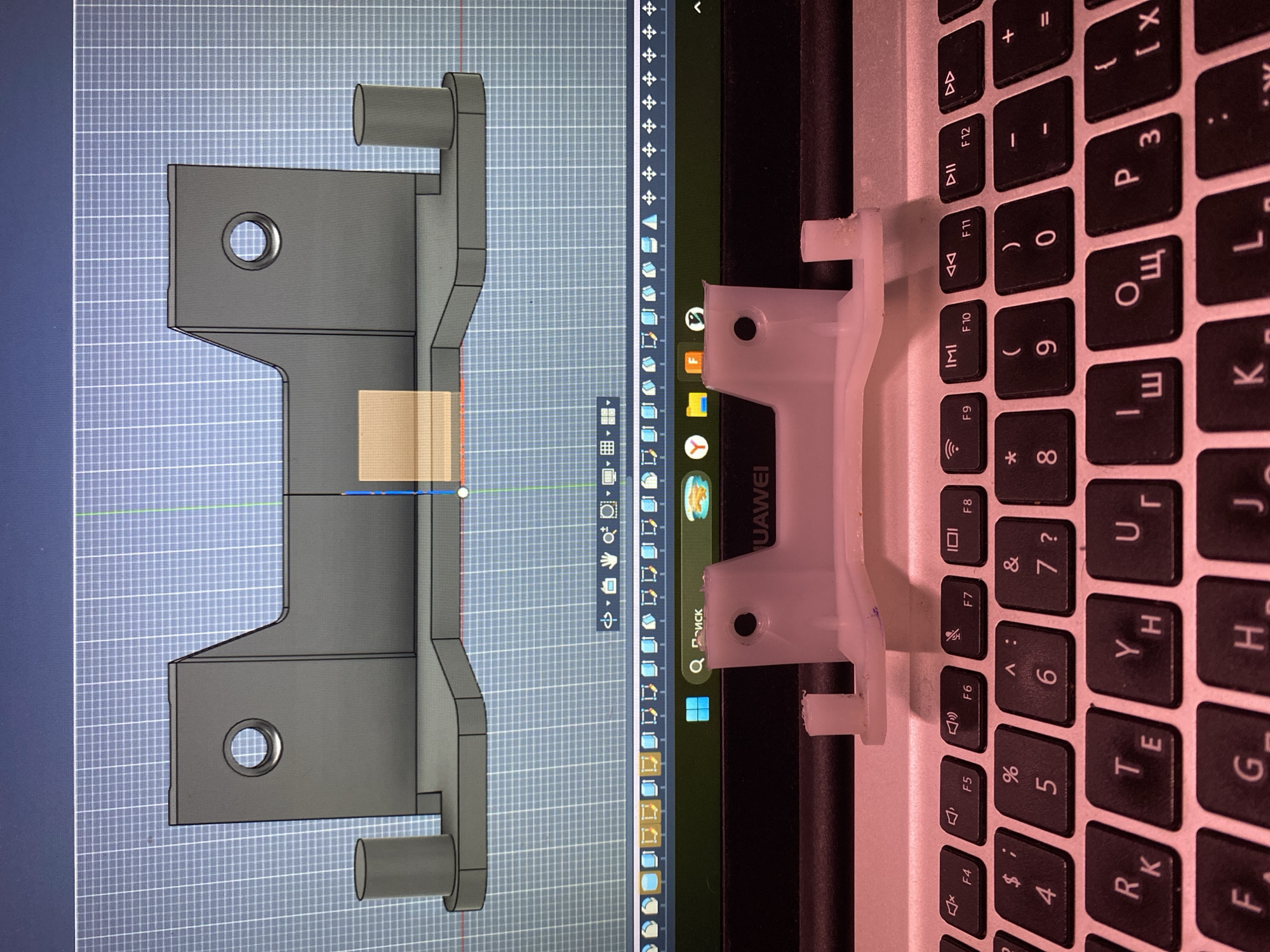 The image shows a comparison between a 3D computer model of a part and a physical 3D printed part placed on a laptop keyboard. The 3D model, rendered in gray, is displayed on a screen with a grid background. To the right, the physical part, made of white plastic, rests on a laptop keyboard, revealing black keys with Cyrillic characters. The laptop appears to be a Huawei brand based on the logo on the screen. Various icons are visible on the laptop screen. There is no indication of a location in Chaikovsky, Russia.