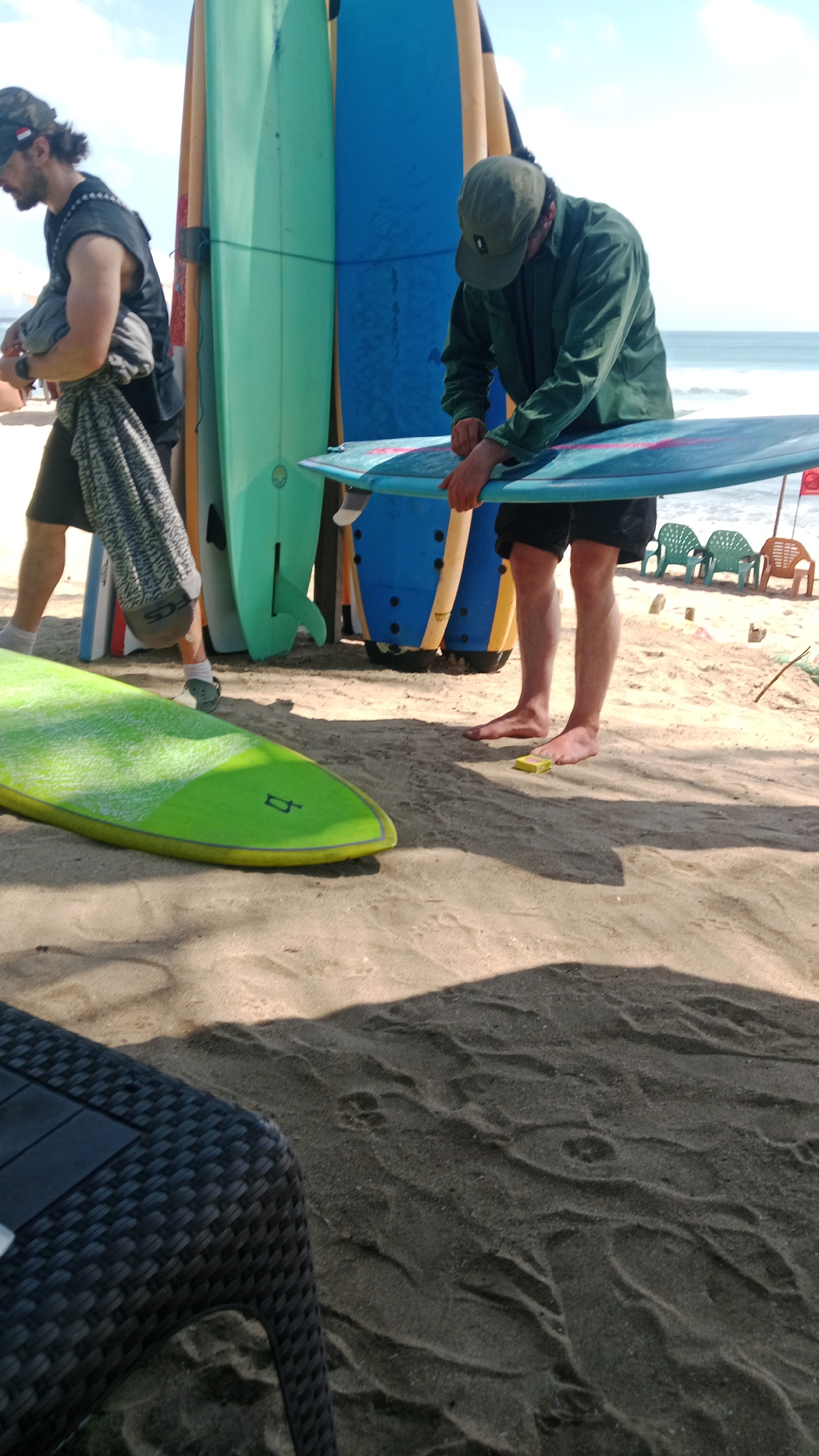 Two men stand on a sandy beach in Kuta, Indonesia, amidst a collection of surfboards. The man on the left, wearing a dark sleeveless shirt, shorts, socks, and a cap, is carrying a folded grey towel or bag. He appears to be walking away from the viewer, his left leg slightly bent. The man on the right, dressed in a green jacket, dark shorts, and a cap, is bent over a blue surfboard, appearing to work on it. His hands are on the board. Several surfboards are propped upright behind them, in various colors including teal, blue, and yellow. A bright green surfboard lies horizontally in the foreground, its nose pointing left. In the background, beach chairs and tables are visible, along with the ocean and sky. The sand is rippled, suggesting the wind or tide.