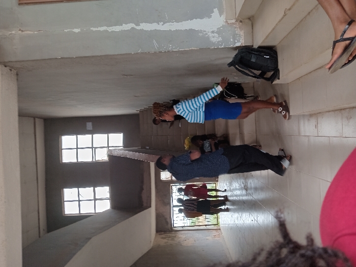 An interior scene captured within a multi-story building in Ogboko, Nigeria, features multiple individuals across various levels. In the immediate foreground, the back of a person's head and right shoulder, wearing a red garment, is partially visible. In the midground, a man in a dark, patterned short-sleeved shirt and black trousers, wearing sandals, stands facing towards the right. Opposite him, a woman with braided hair, wearing a horizontal blue and white striped long-sleeved top and a blue skirt, stands facing left, appearing to interact with the man. Her hands are slightly raised. A black backpack rests on the stairs beside her. Another person is partially visible behind the man, seated on a lower step of a concrete staircase that ascends to the upper levels.
Further into the background, near an open entranceway, a group of approximately three to four individuals are standing. One individual wears a red dress, another a patterned top and dark skirt. The building exhibits unfinished concrete walls, some showing signs of flaking paint or plaster. Two large windows with grid patterns are visible on the upper wall, allowing bright light into the space. The floor is covered with light-colored tiles. A concrete staircase with a dark railing ascends from the ground floor to the upper stories, with a landing visible halfway up. The overall environment suggests an institutional or commercial building, possibly undergoing construction or in a state of wear.