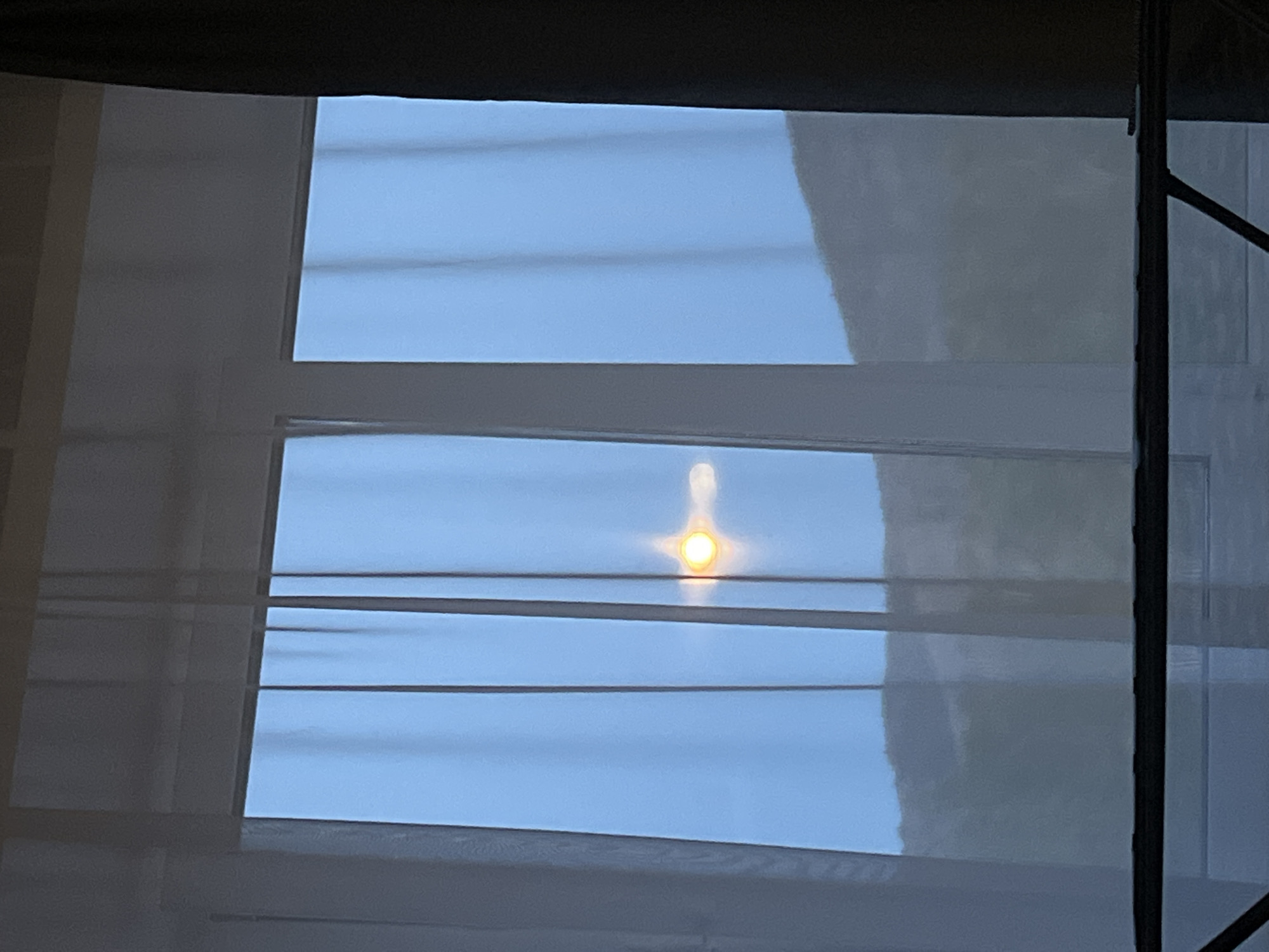 The image captures an outdoor scene viewed through a window partially obscured by light-colored, possibly sheer, horizontal blinds or curtains. The window features a white frame, dividing it into multiple panes through which a clear blue sky is visible. A prominent, bright yellow-orange light source, likely the sun, is centered in one of the middle window sections, emitting a distinct halo and a vertical streak of light extending upwards, possibly a lens flare or reflection.
The translucent blinds create horizontal bands across the view, allowing the blue sky to filter through while diffusing the light. The overall lighting suggests it is daytime with clear weather, as no clouds are visible. On the far right side of the image, a dark, thin vertical bar or post is partially visible, which could be part of the window's exterior structure or a nearby object. The interior space from which the photo is taken appears darker in comparison, indicated by the shadowed upper edge. No people are present, no specific activities are taking place, and there is no visible text.