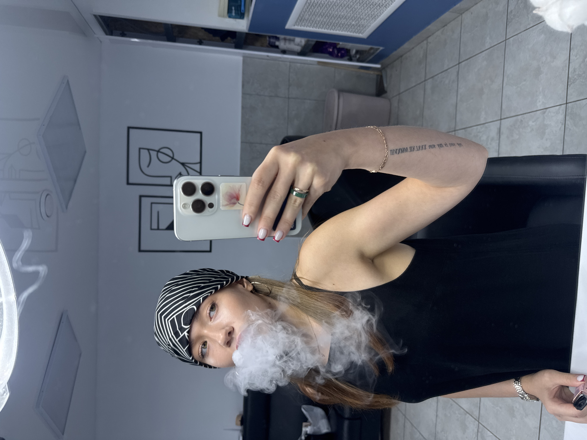 A young woman with long, brown hair, wearing a black tank top and a black and white patterned bandana, is taking a selfie in a brightly lit room. She is exhaling a cloud of white smoke, likely from an electronic cigarette, which obscures the lower half of her face. Her right hand holds a white smartphone, and on her left wrist, she wears a silver watch. She has a gold bracelet on her right wrist and a ring with a green stone on her right index finger. In the background, a mirror reflects the room, revealing a tiled floor, a black couch, a trash can, and minimalist artwork on the wall. The room appears to be a salon or personal studio, possibly a beauty salon, given the presence of shelving units with bottles and containers in the background. There is no specific identifiable information suggesting the location is in the city of Chaikovsky, Russia.