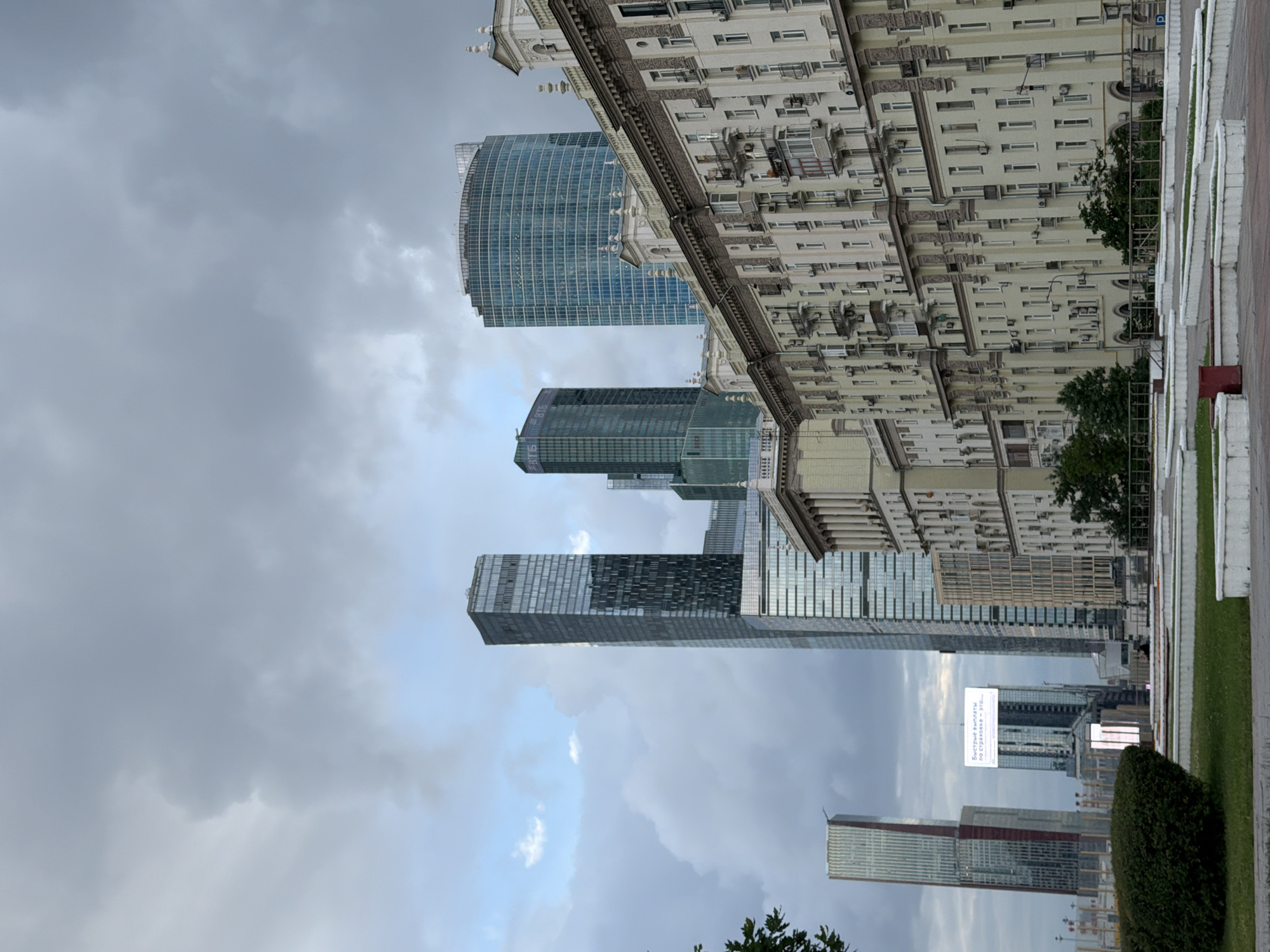 This media file depicts an urban scene in Moscow, Russia, featuring a prominent display of modern skyscrapers and an older, ornate building. The skyscrapers are characterized by their glass facades and sleek, vertical lines, with one particularly notable curved blue glass building. In contrast, the older building exhibits classic architectural elements, with intricate detailing and numerous windows.
The overall scene is dominated by a cloudy, overcast sky, suggesting either early morning or late afternoon, with the weather appearing somber. The perspective is from a slightly elevated viewpoint, looking upwards at the towering structures. Greenery, including trees and manicured bushes, is visible in the foreground and interspersed between buildings, indicating a park or landscaped area within the city.
A notable detail is a sign on a smaller building in the mid-ground, displaying Cyrillic text. The text reads "Быстрые билеты по страховке - это..." which translates to "Fast tickets for insurance - this is...". This suggests the presence of commercial activity within the urban landscape. The composition emphasizes the juxtaposition of modern and historical architecture within a bustling city environment. No people are directly visible in the frame, and the overall mood conveyed is one of architectural grandeur under a subdued sky.