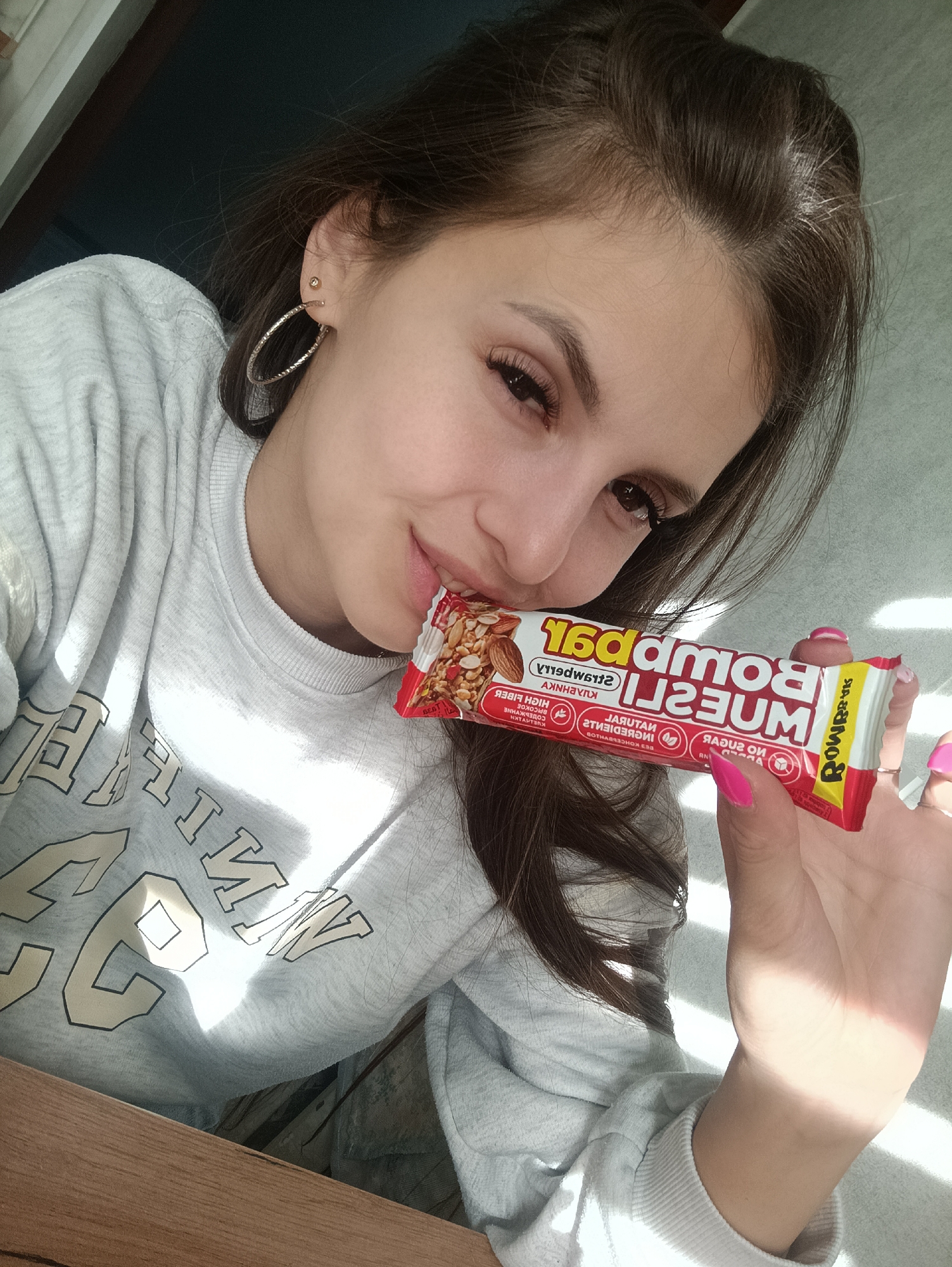 A woman is holding a Bombbar Muesli bar in her right hand, with the end of the bar between her lips. She is wearing a light grey sweatshirt with the letters "WIN" partially visible in white and black. She has long brown hair pulled back and is wearing hoop earrings. Her fingernails are painted pink. The background appears to be a neutral colored wall, and a corner of a brown wooden surface is visible. The city and location are unknown.
